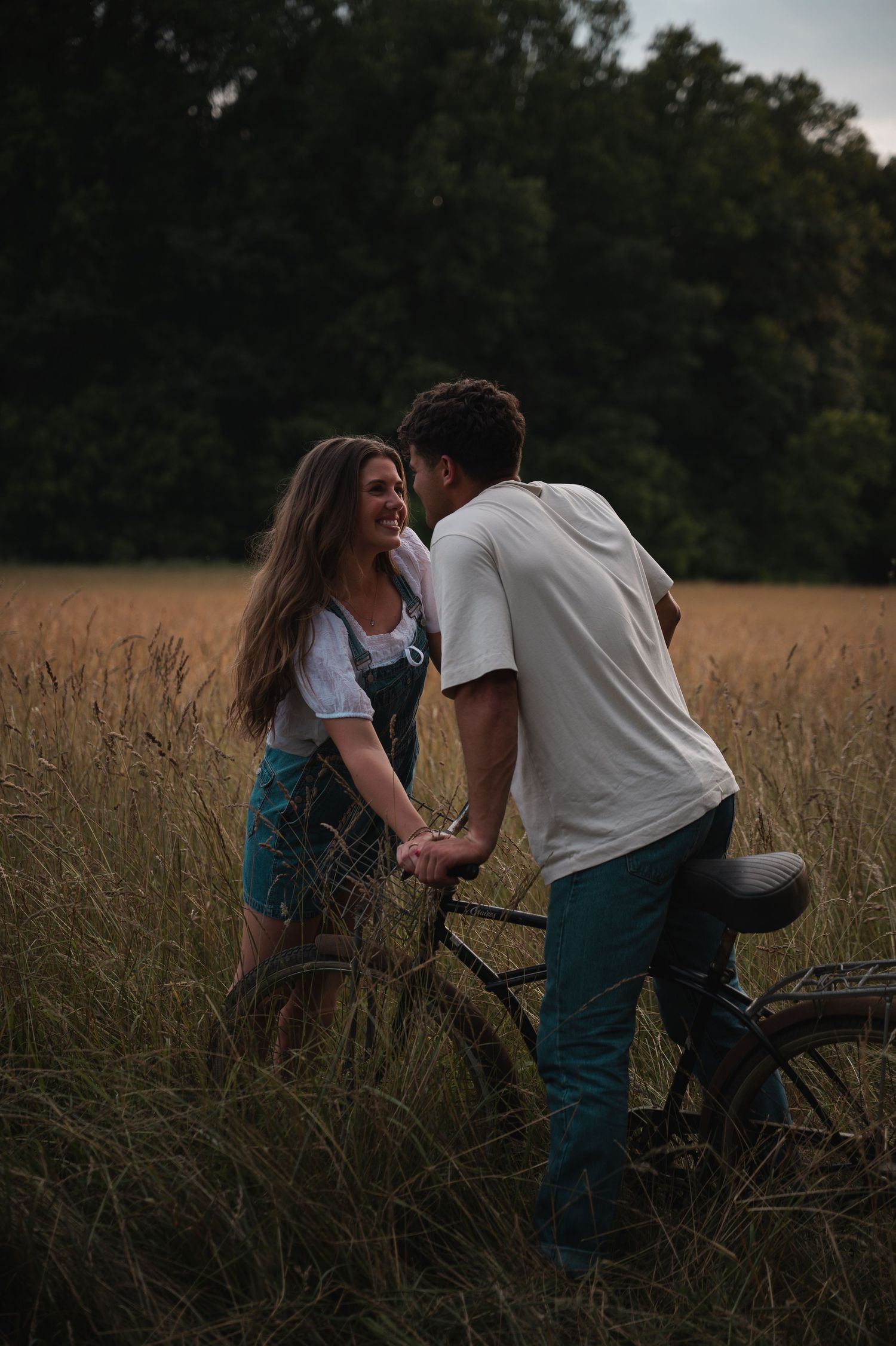 A couple holds hands and laughs together beside a vintage bicycle in a golden field at dusk.