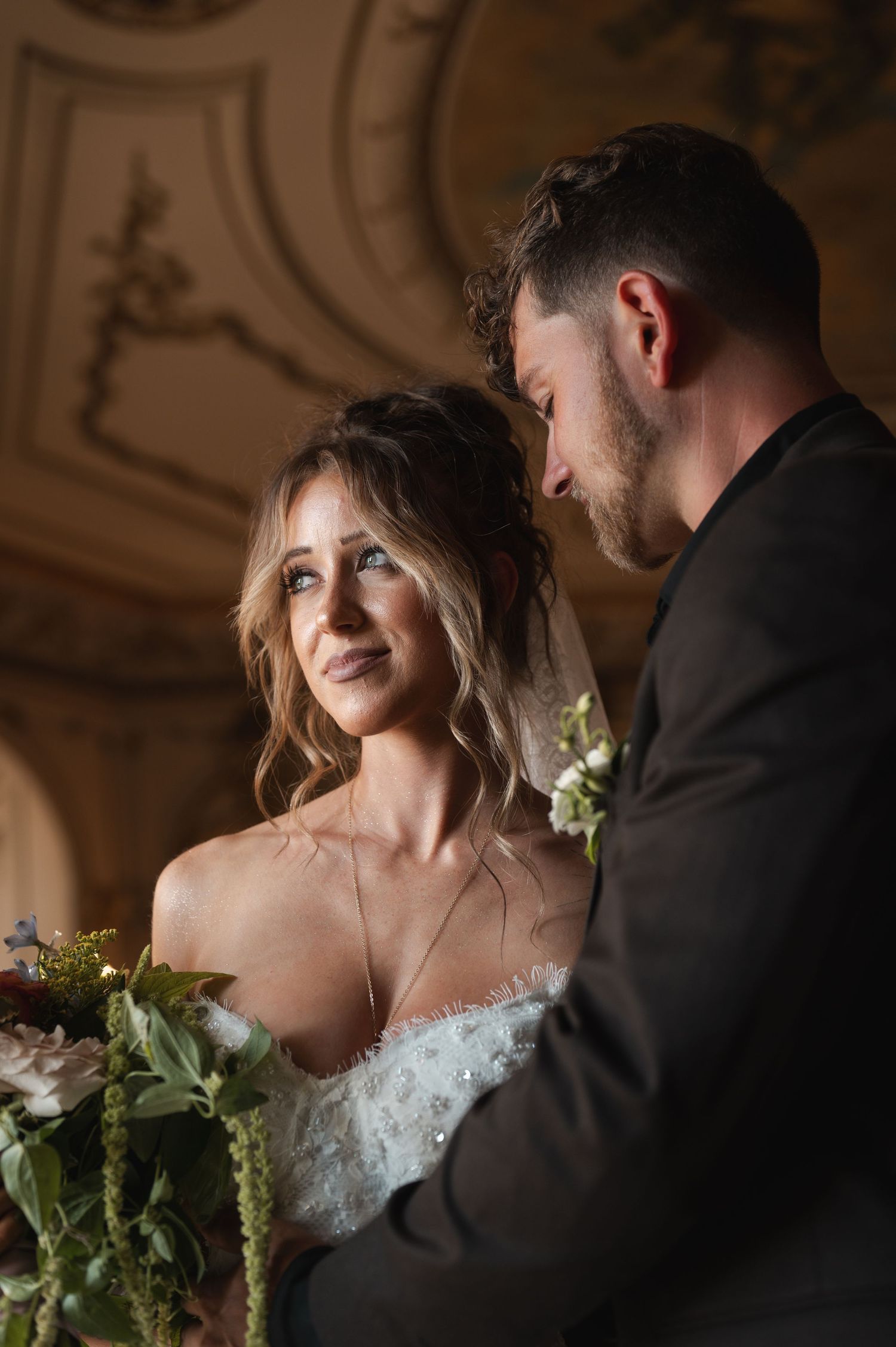 Bride in white gown holds floral bouquet while groom in dark suit leans in close during romantic wedding ceremony.