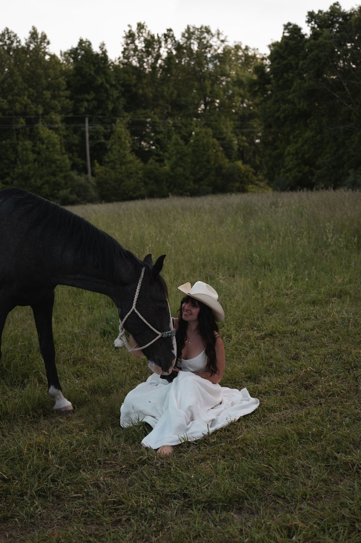 Woman in white dress and cowboy hat sits in green field beside a black horse at dusk.