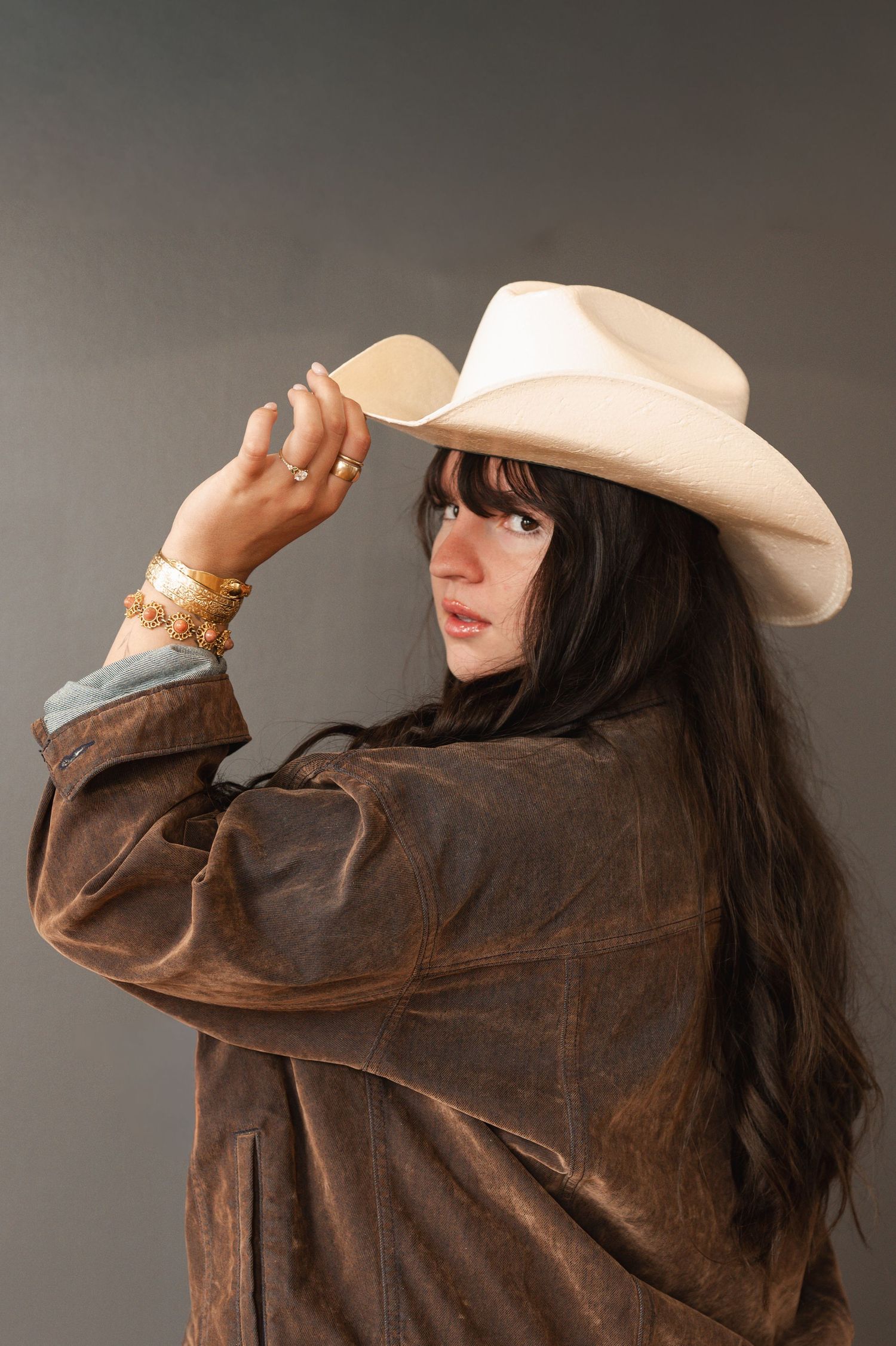 Woman in brown leather jacket and white cowboy hat adjusting brim, wearing gold bracelets and rings.