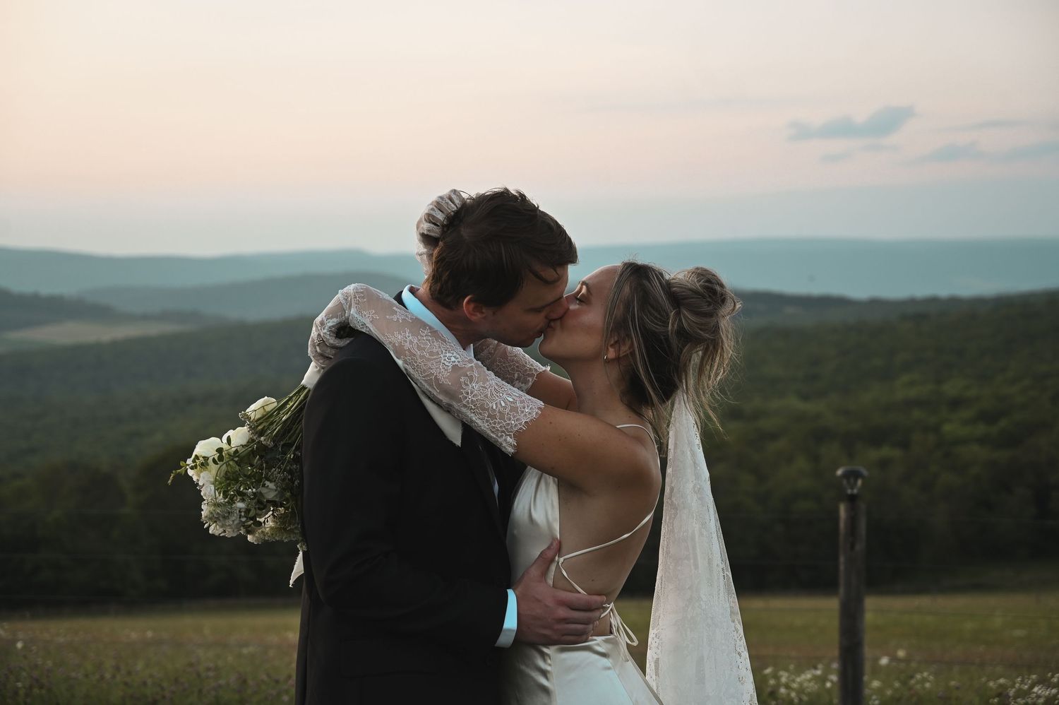 A bride and groom share a romantic kiss at sunset with scenic mountain views in the background on their wedding day.