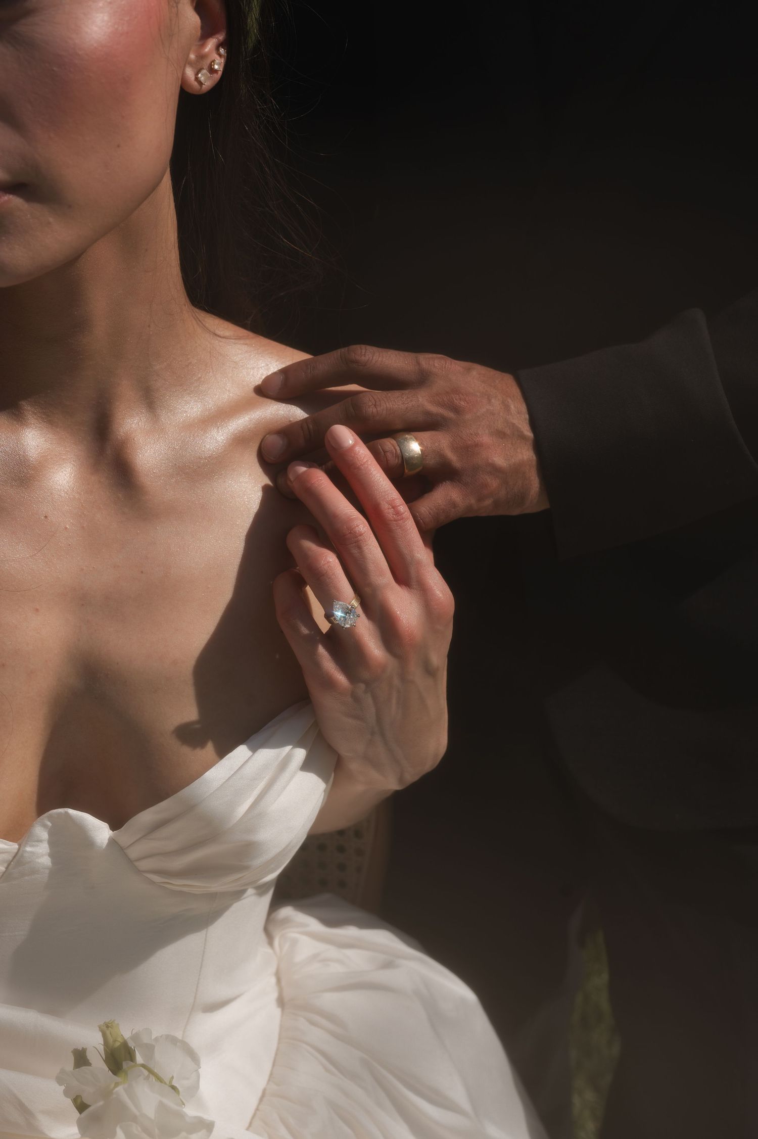 Close-up of bride in white gown holding groom's hand, showcasing elegant engagement ring in soft natural light.