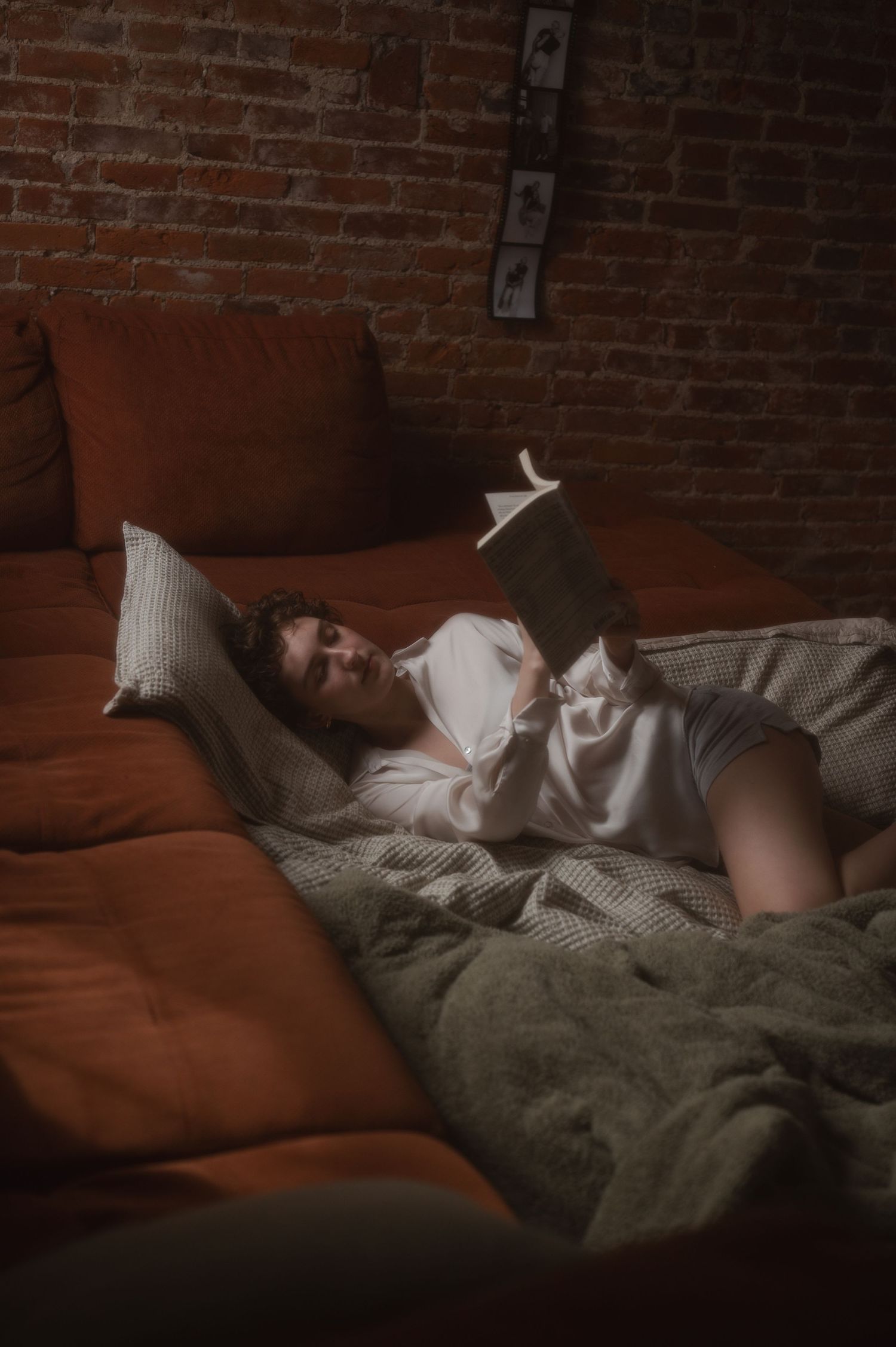A woman relaxes in bed reading a book, surrounded by orange pillows and green bedding against a brick wall backdrop.