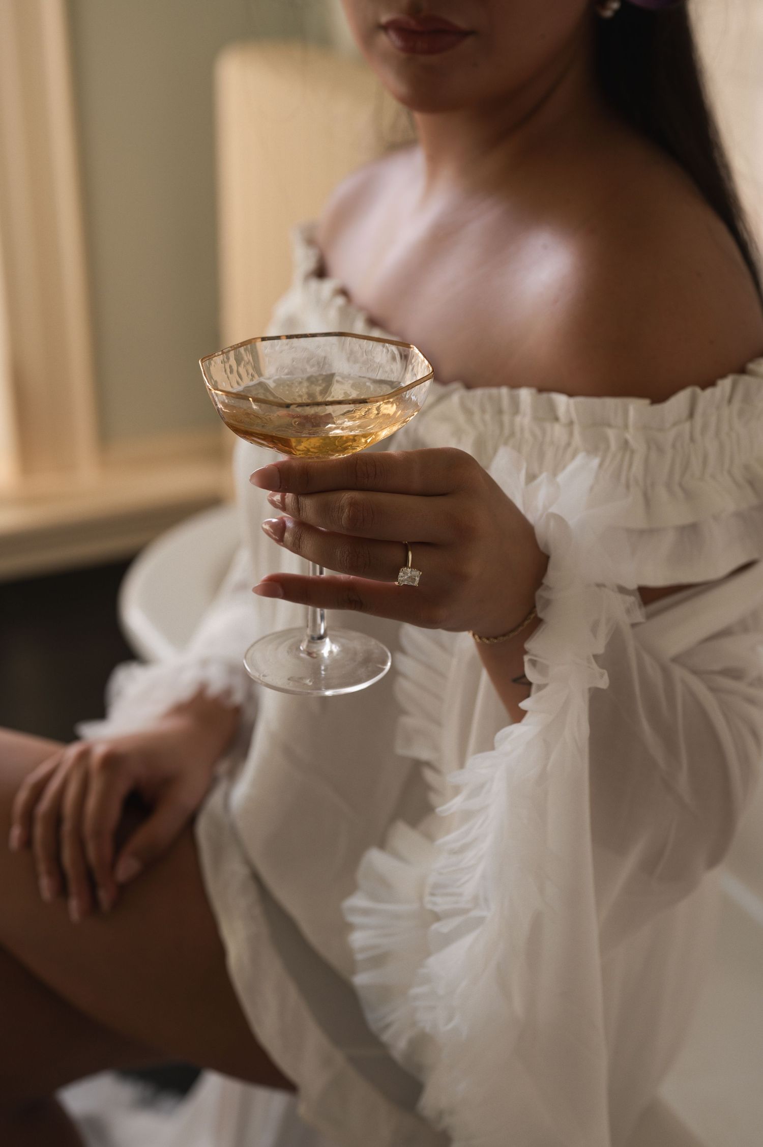 Woman in white off-shoulder dress holding a coupe cocktail glass with a diamond engagement ring on her finger.