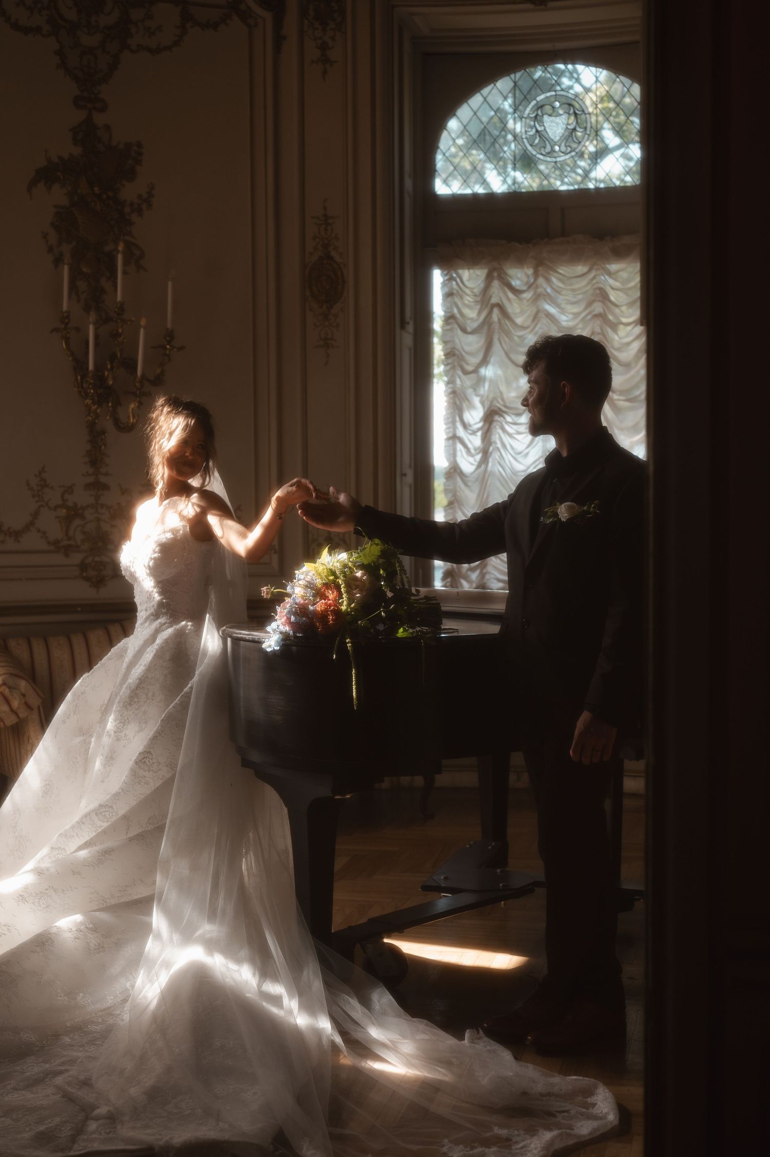 Bride and groom dancing by a grand piano in an elegant sunlit room with ornate decor and a floral bouquet.