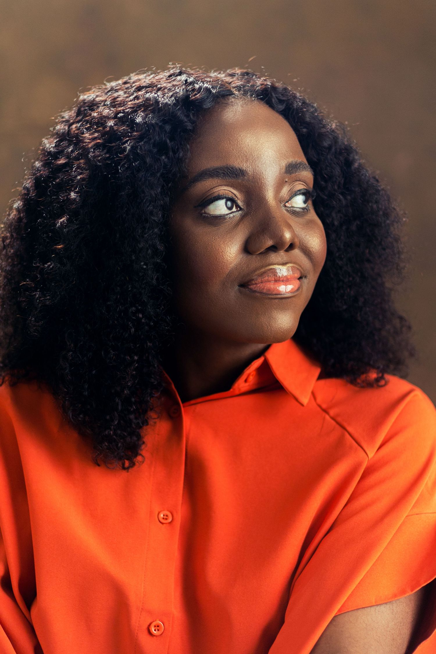 Close-up portrait of a person in a vibrant orange shirt with natural curly hair against a brown background.