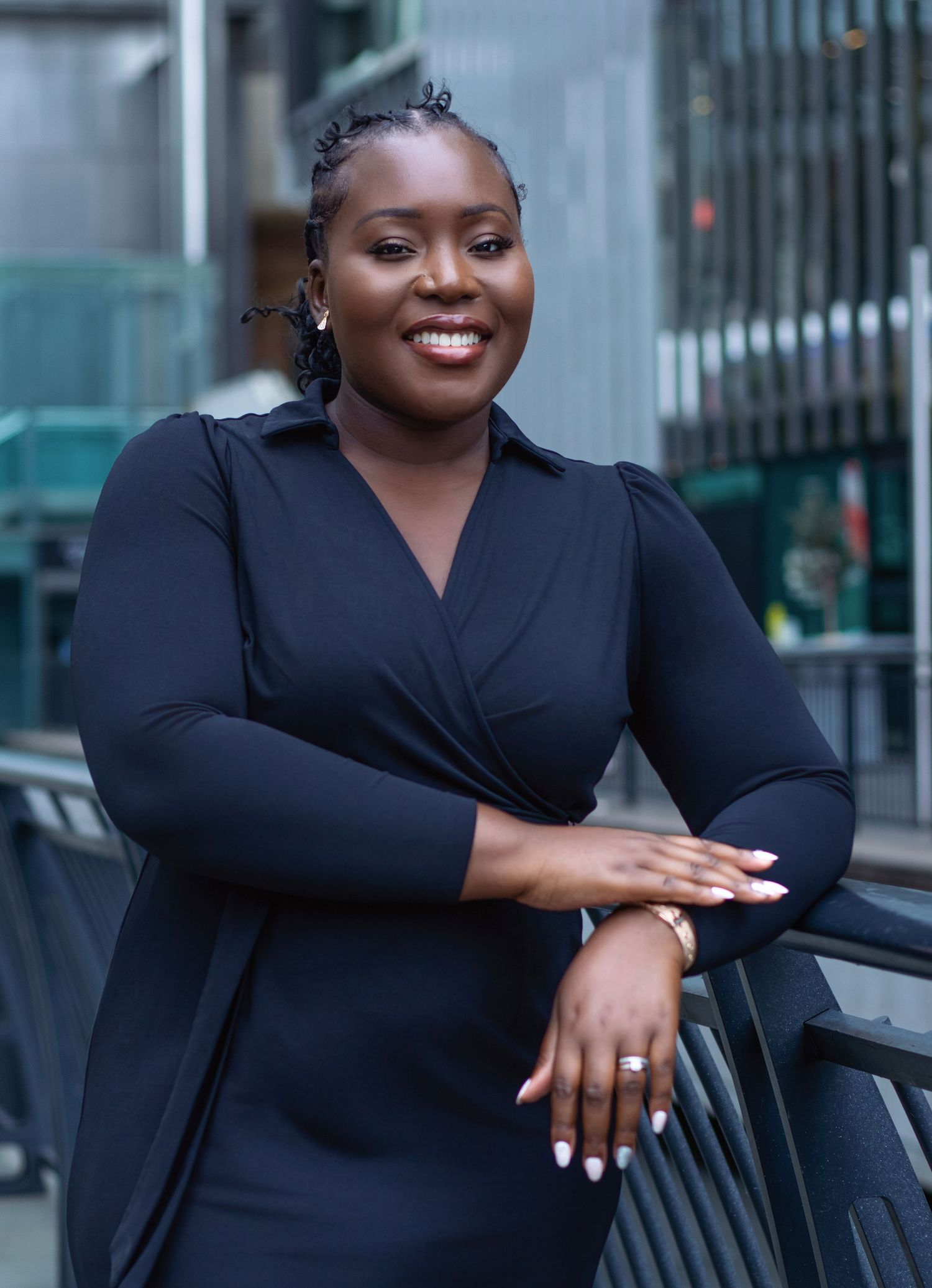 A smiling woman in a black wrap dress leans on a railing in an urban setting, exuding confidence.