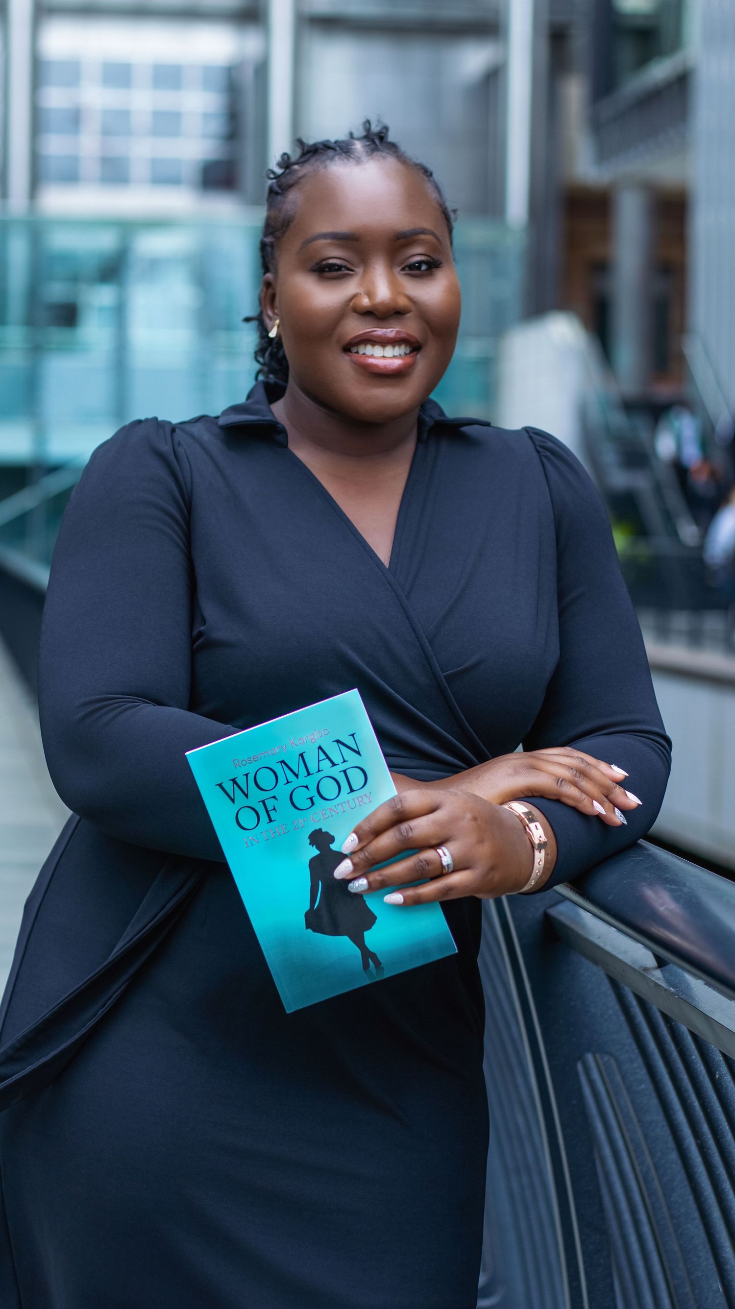 Woman in black dress smiling and holding a teal book titled Woman of God in a modern building.