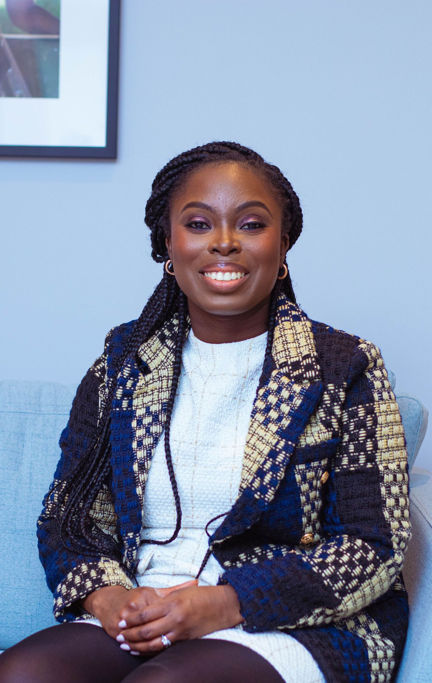 Seated figure wearing a white dress and patterned navy and gold blazer smiles warmly on a blue couch.