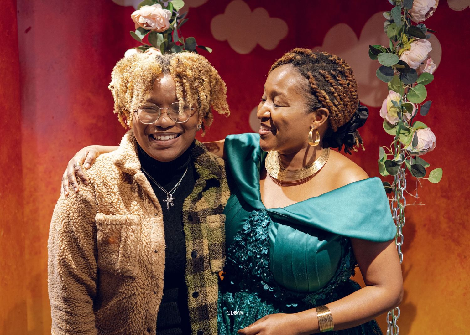 Two women smiling and embracing in front of a red wall decorated with pink flowers and a swing.