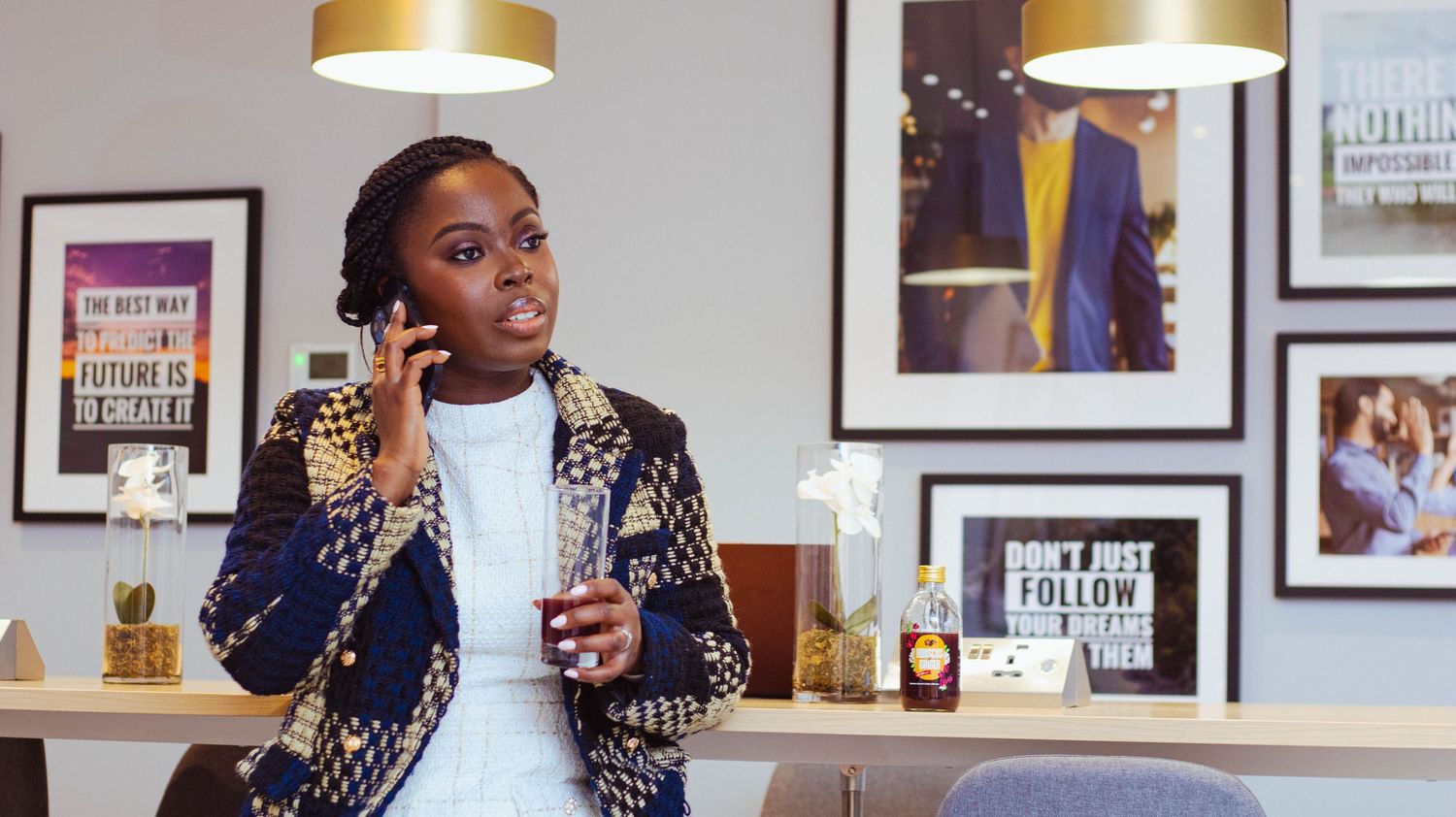 Person in a patterned jacket speaks on phone while working in modern office space with framed artwork and pendant lights.
