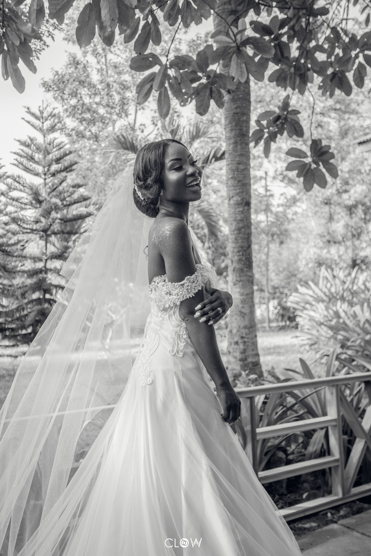 A bride in a stunning white off-shoulder wedding gown and long veil poses gracefully in a garden setting.
