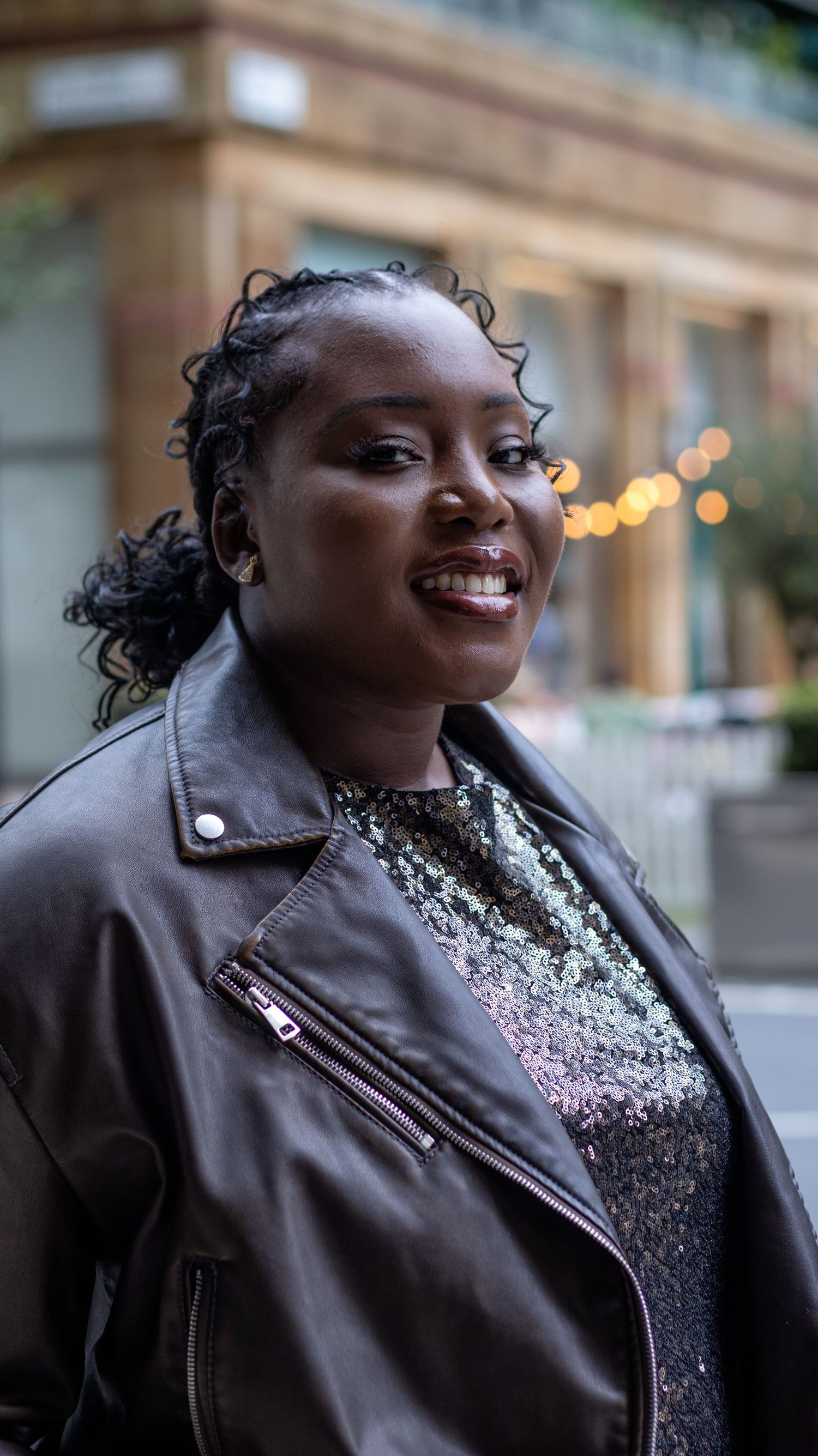 A person wearing a brown leather jacket and sparkly top smiles warmly in front of blurred city lights at dusk.