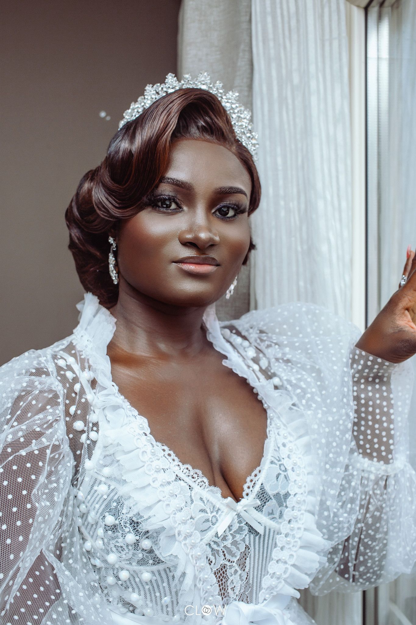 Elegant bride in white lace gown and crystal tiara poses gracefully near a window in soft natural light.