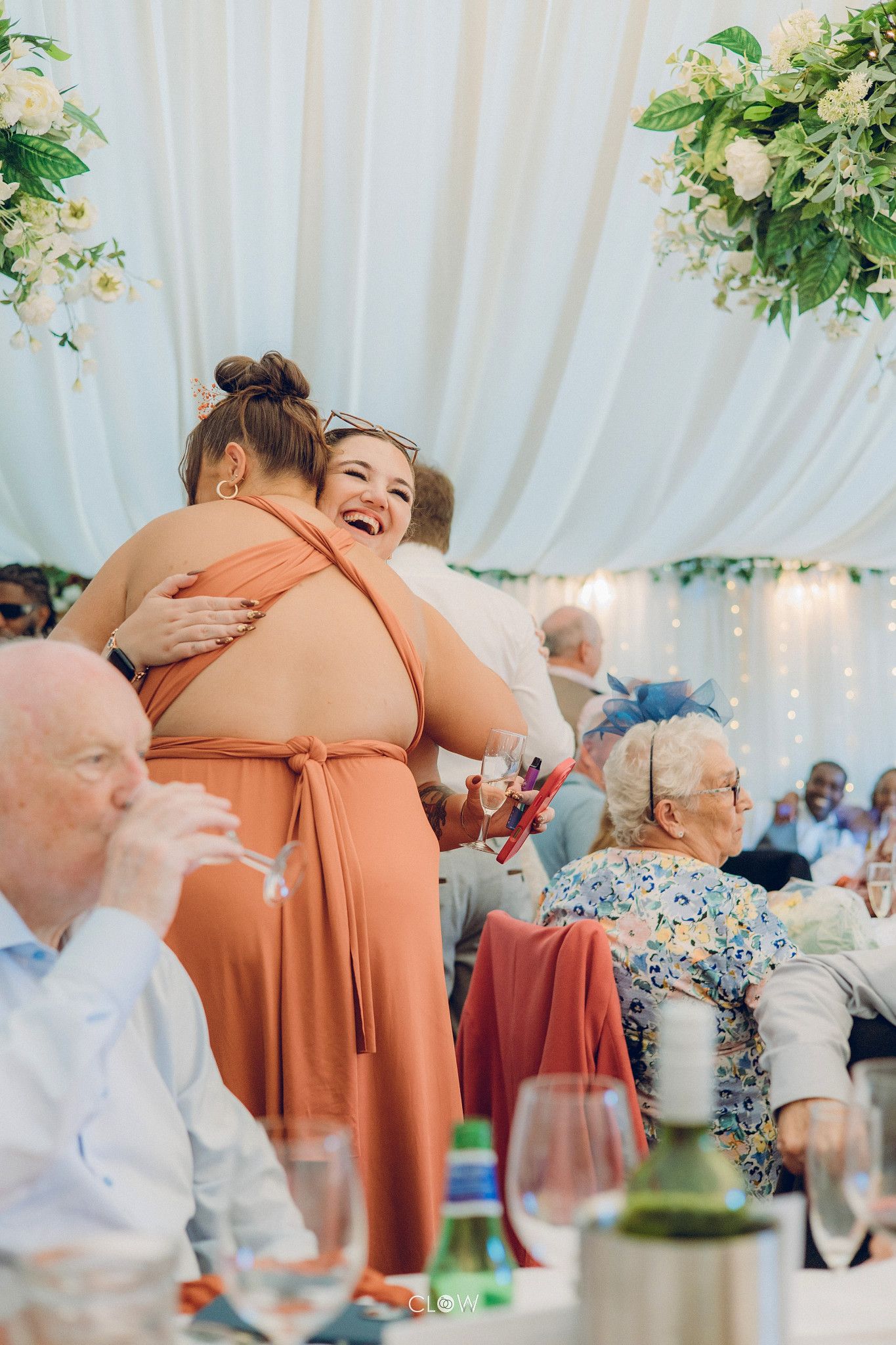 Two women share a joyful hug at an elegant wedding reception under a white draped marquee with floral decorations.