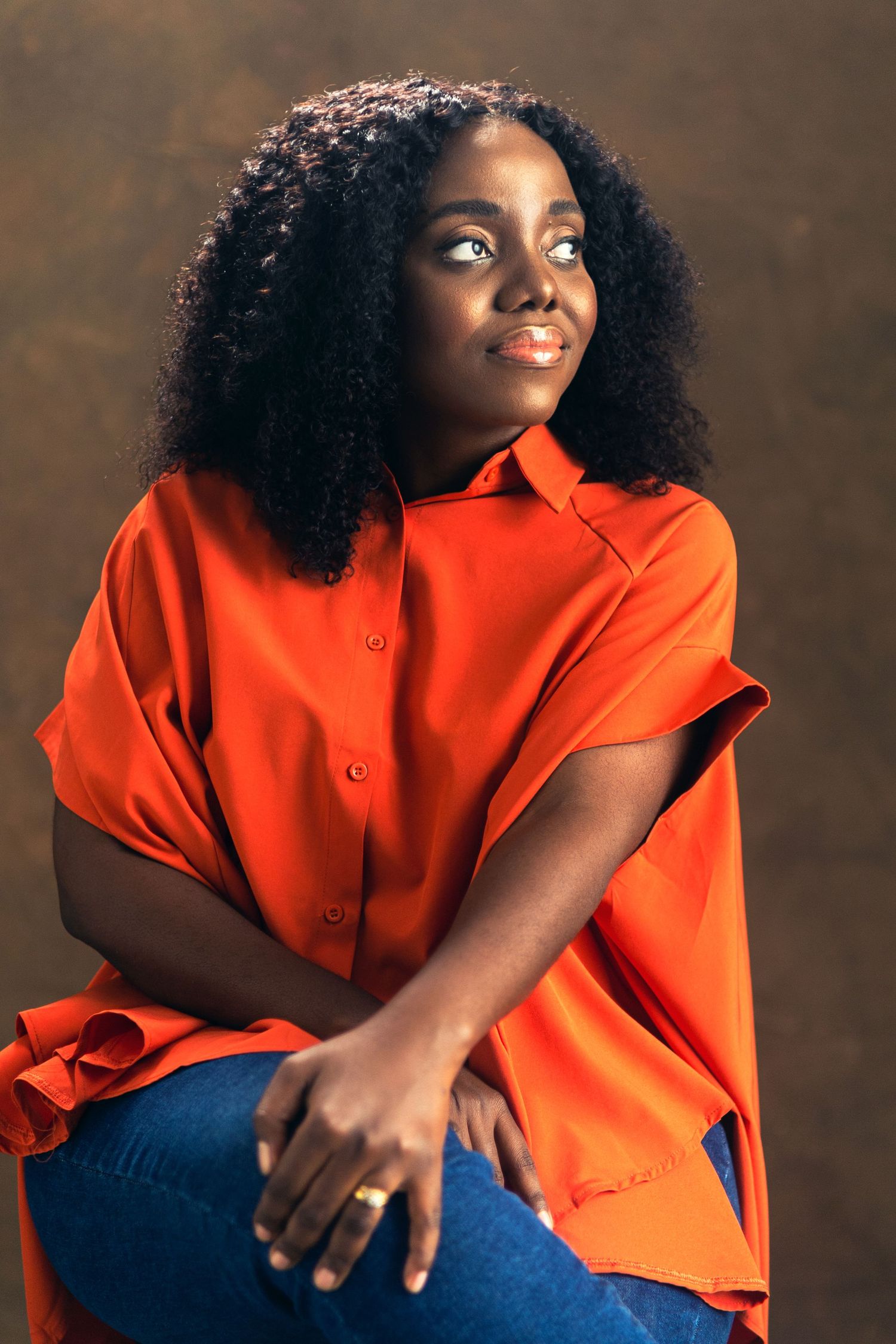 Portrait of a person in a bright orange button-down shirt and blue jeans looking off-camera against a brown backdrop.