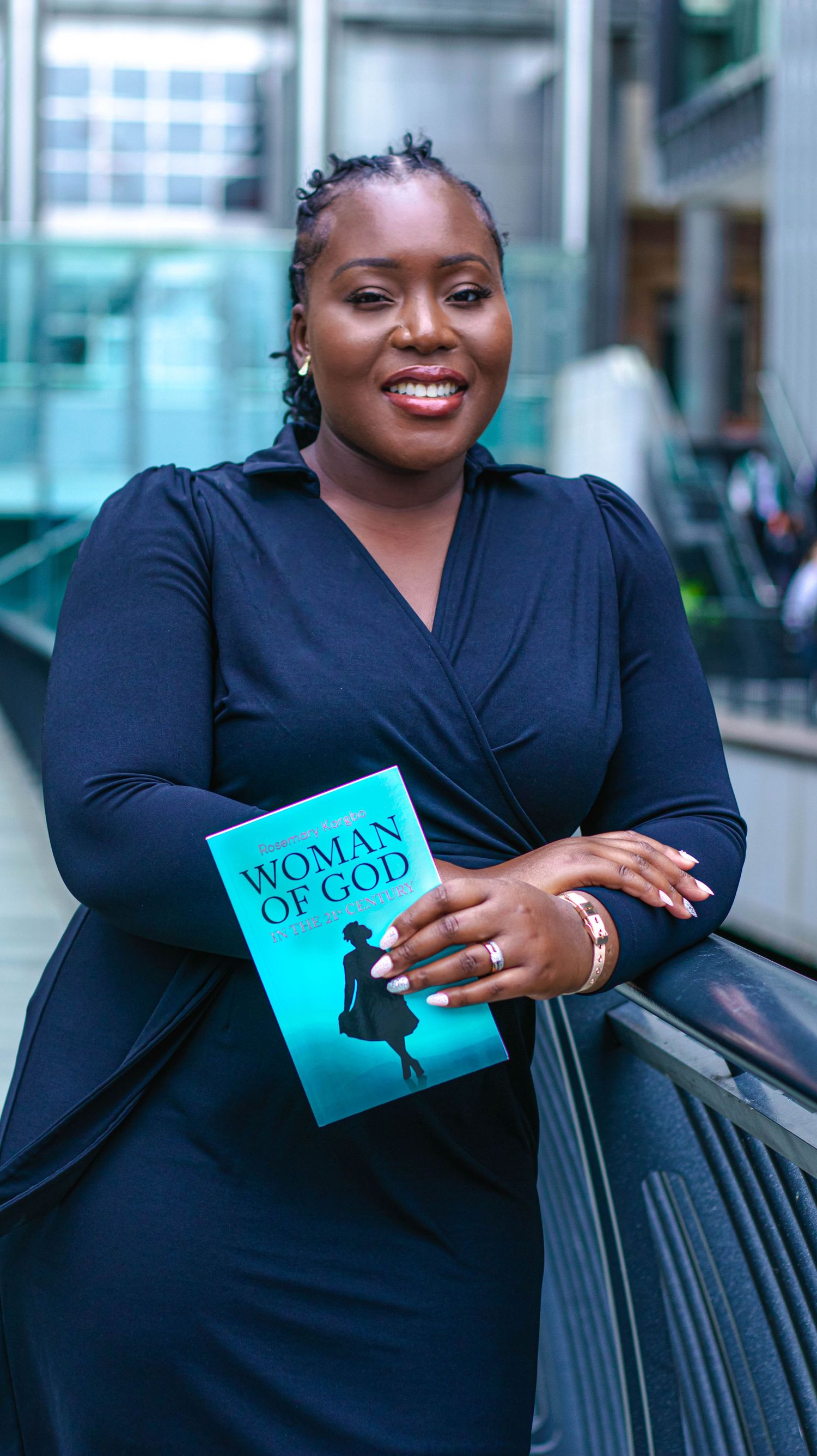 Smiling woman in navy dress holds turquoise book while posing professionally outside modern building.