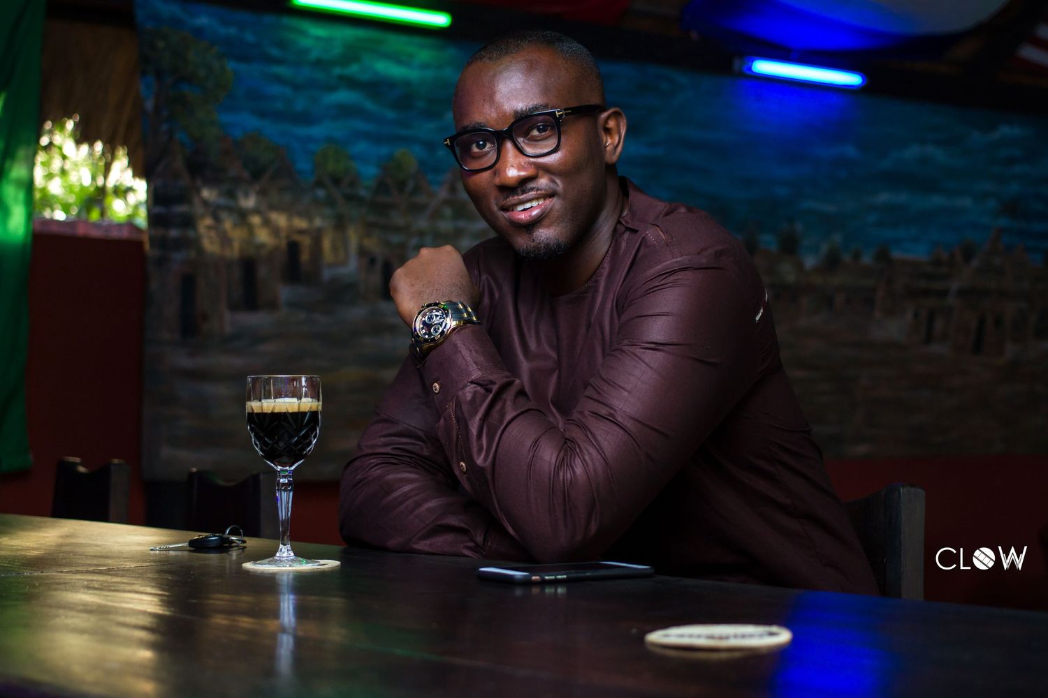 Person wearing glasses and burgundy jacket sits at dimly lit bar with neon blue and green lighting in background.