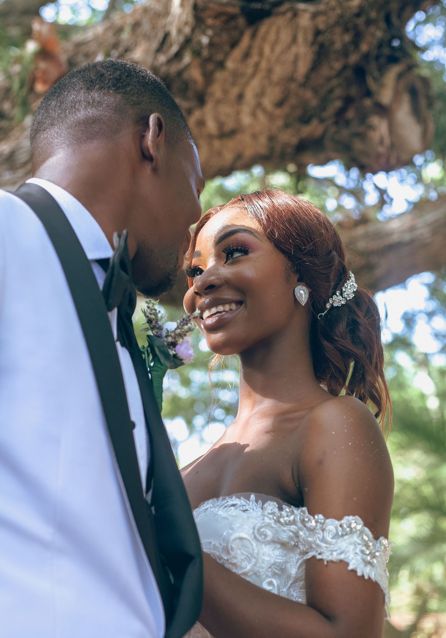 A joyful bride in an off-shoulder lace gown smiles at her groom in a white suit during an outdoor wedding ceremony.