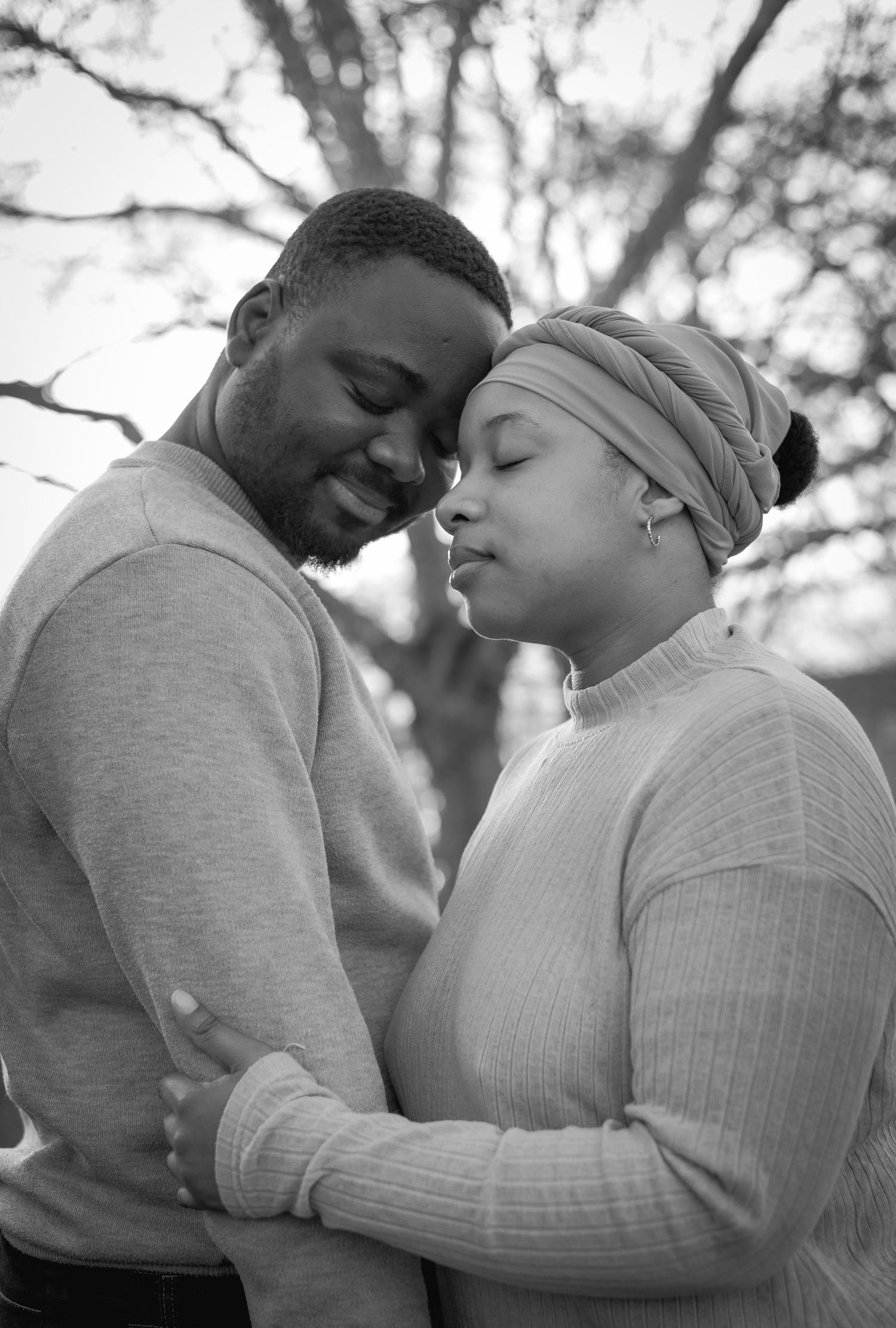 Black and white photo of a couple embracing tenderly, foreheads touching, outdoors with bare trees in background.