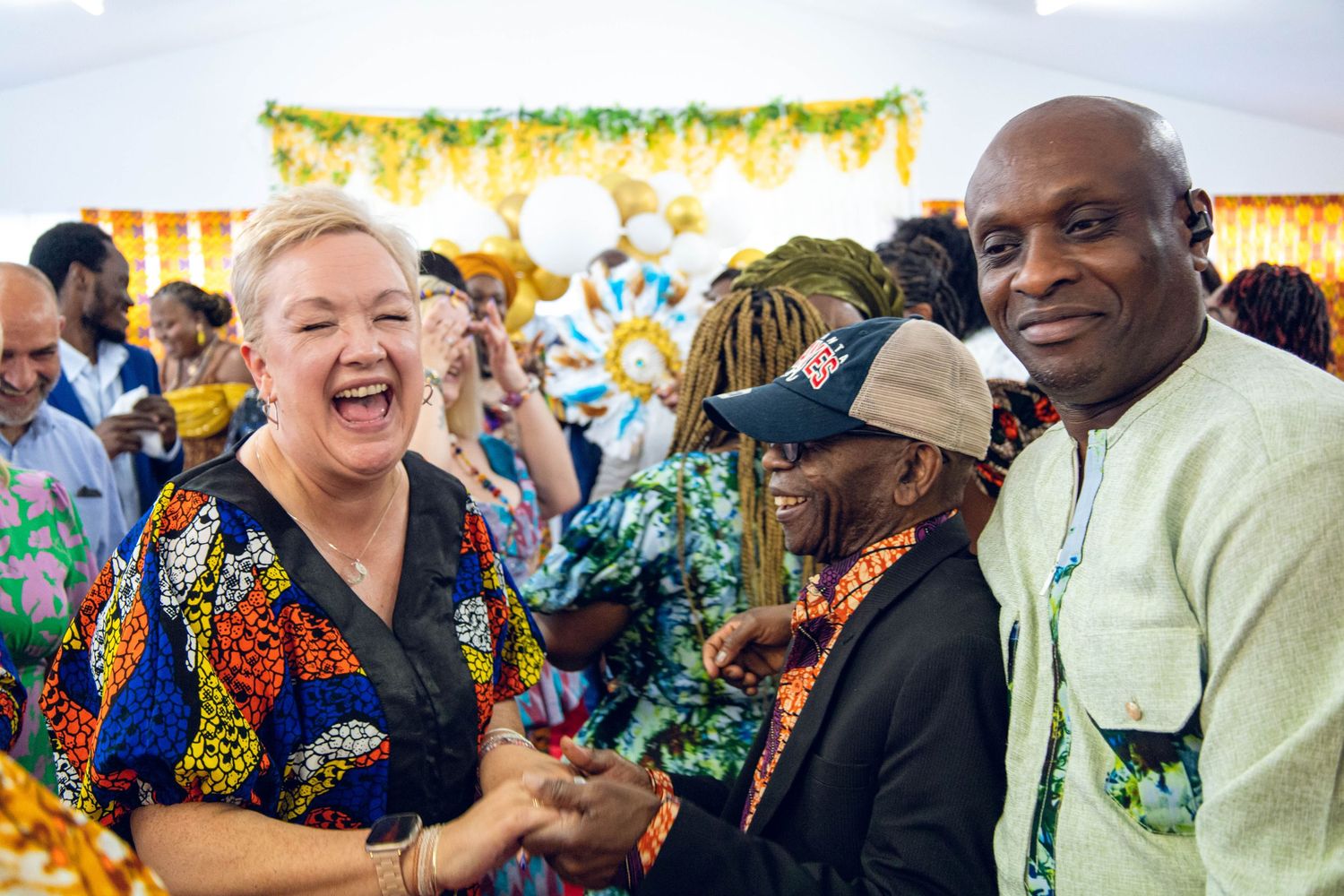 A group of people wearing vibrant colorful clothing laugh and smile together at an outdoor festival celebration.