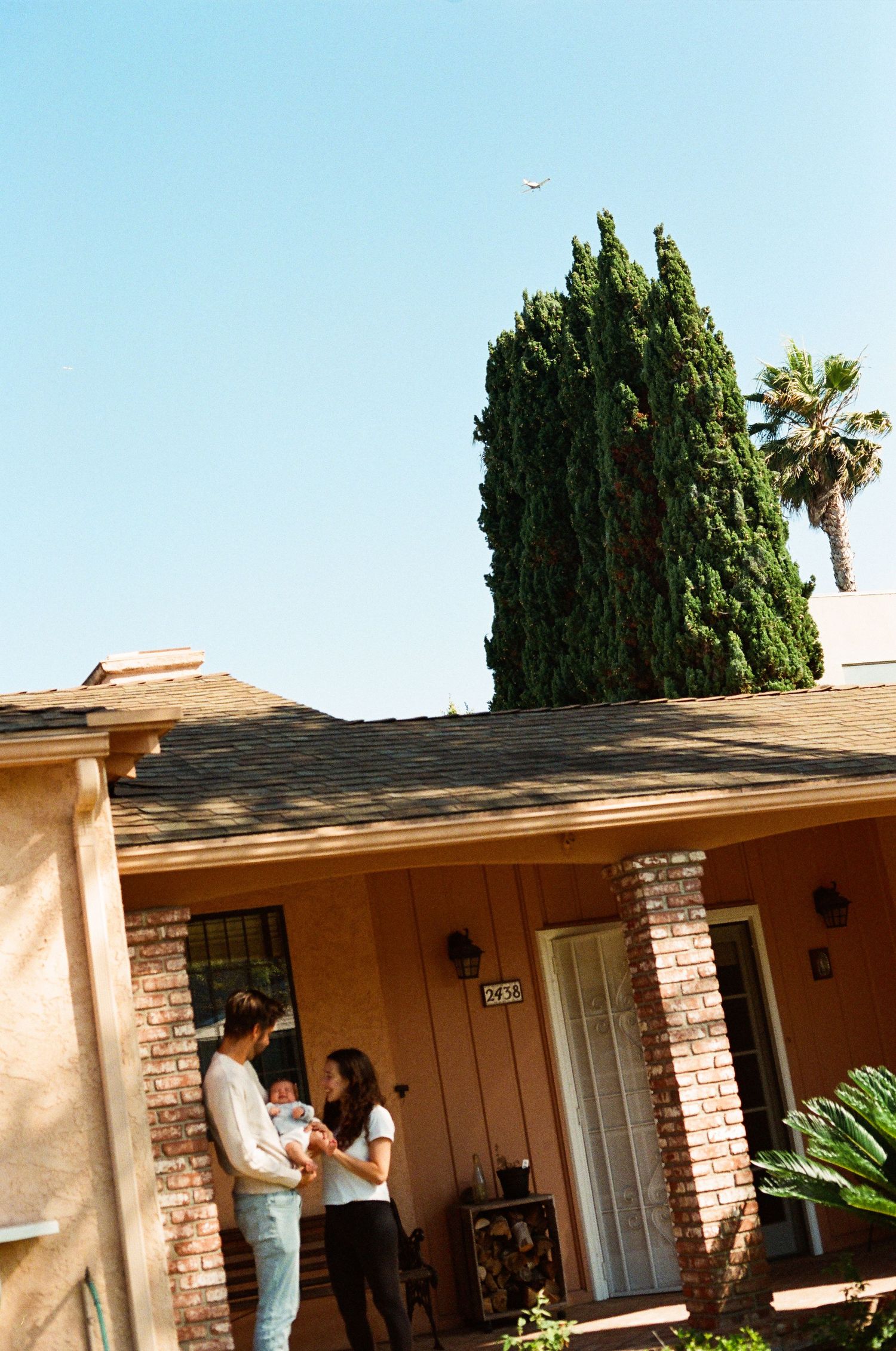Two women holding newborn baby stand on porch of suburban brick home under blue sky.