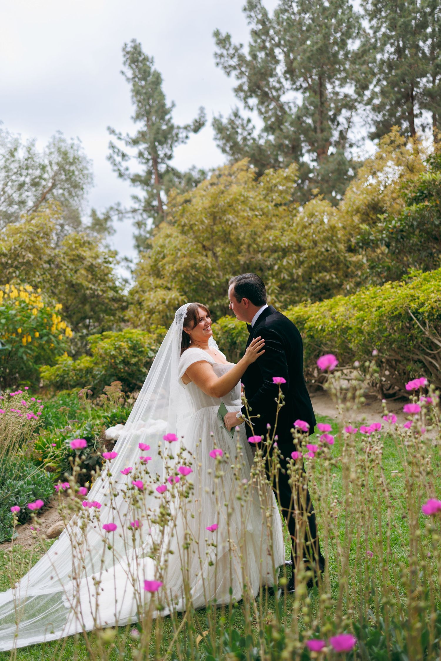 A bride and groom embrace tenderly among blooming pink flowers in a lush garden with tall trees in the background.