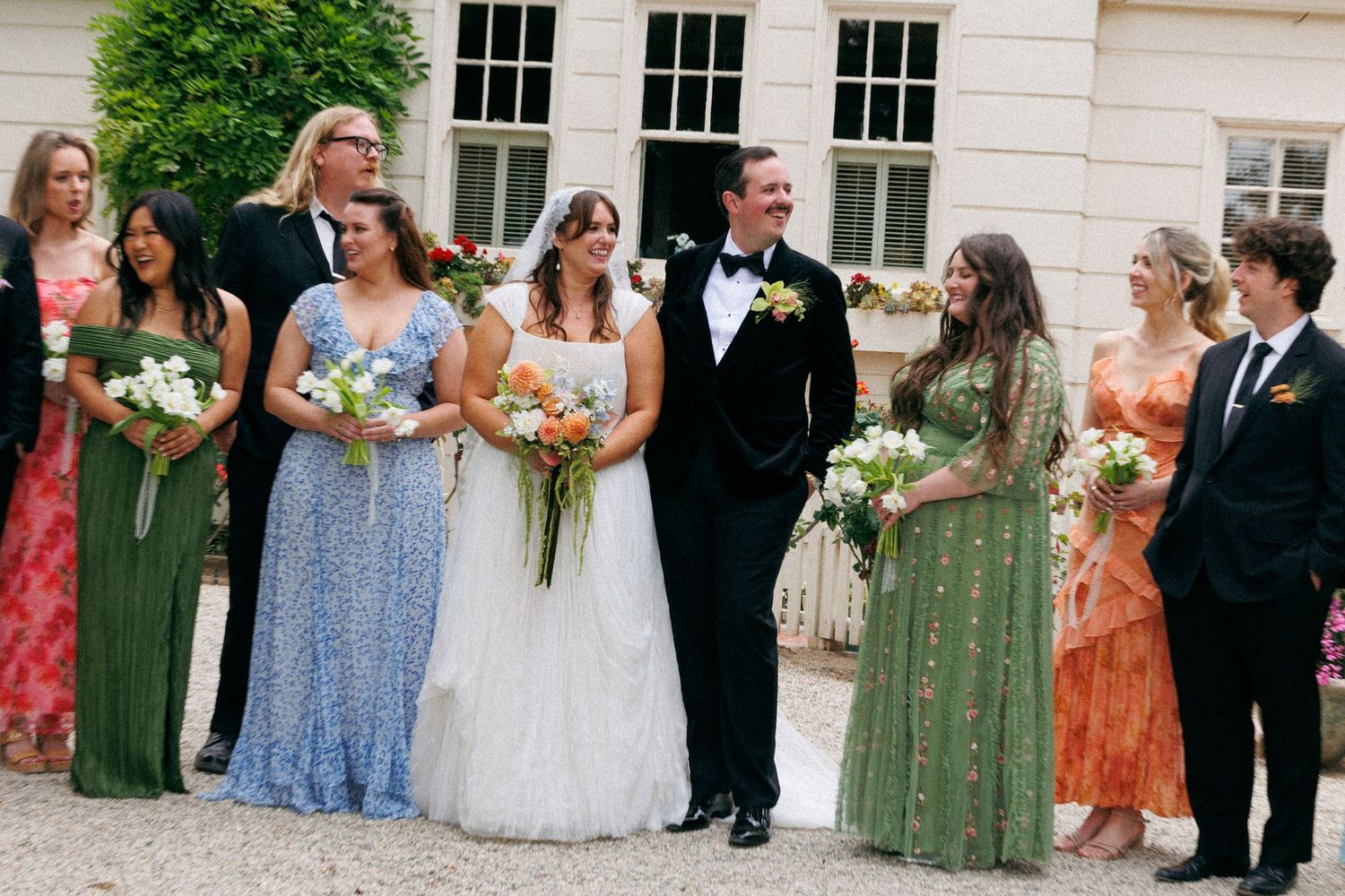 Wedding party poses outside chateau with bridesmaids in colorful green and floral dresses flanking bride and tuxedoed groom.
