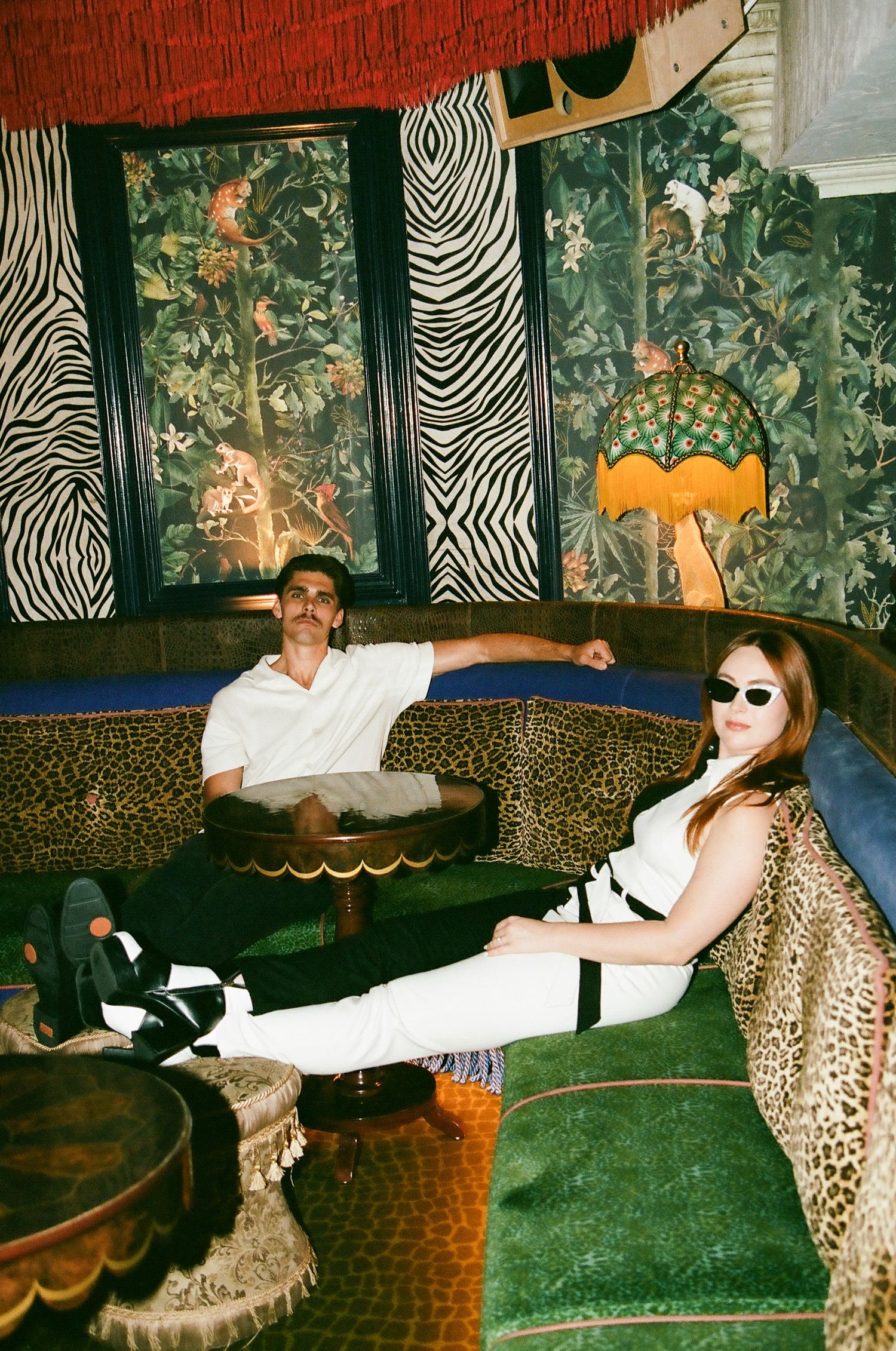 Couple relaxing on leopard print sofa in eclectic bar with zebra wallpaper and jungle murals.