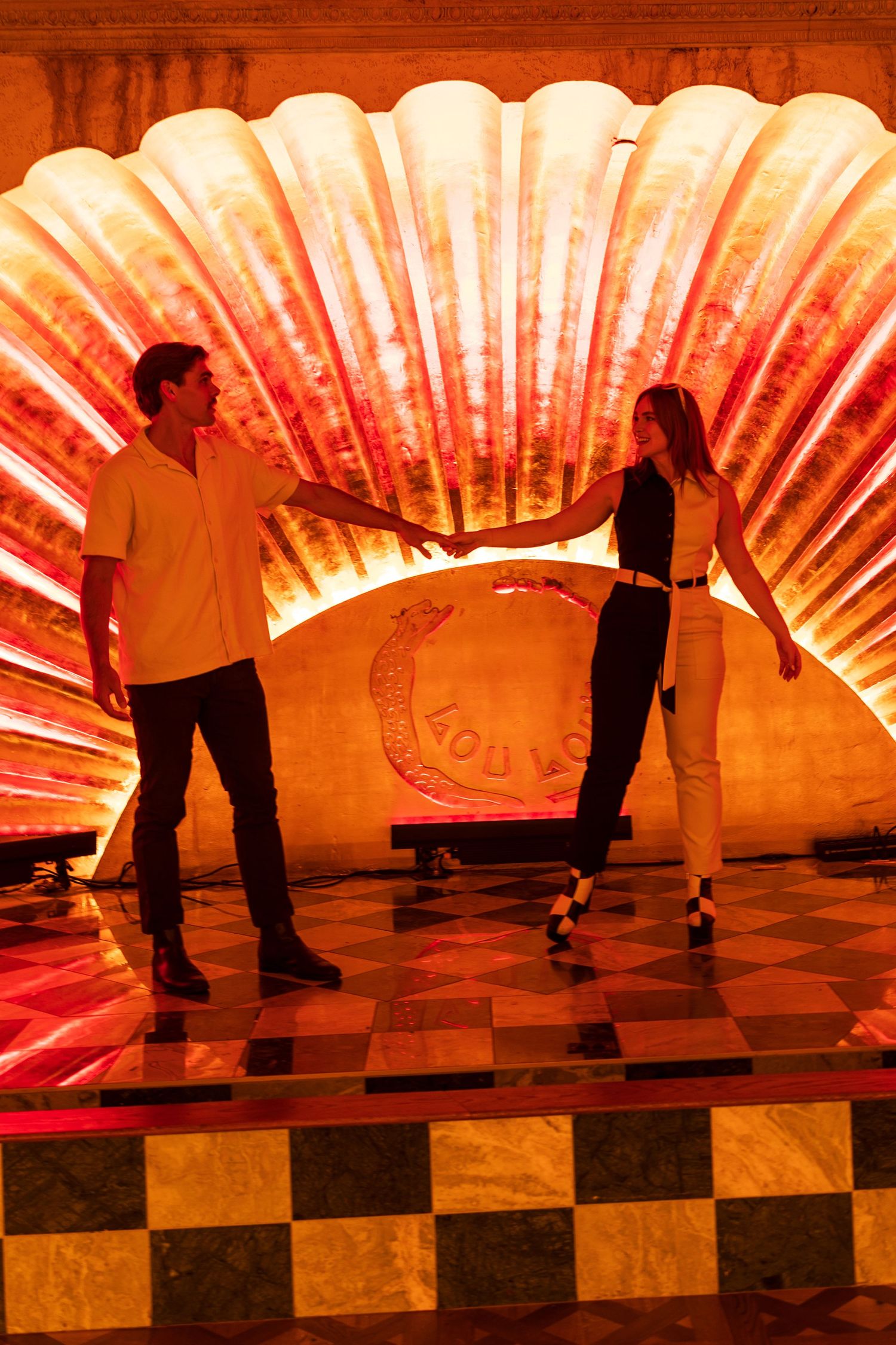 Couple holding hands on checkered floor in front of glowing illuminated shell wall sculpture.