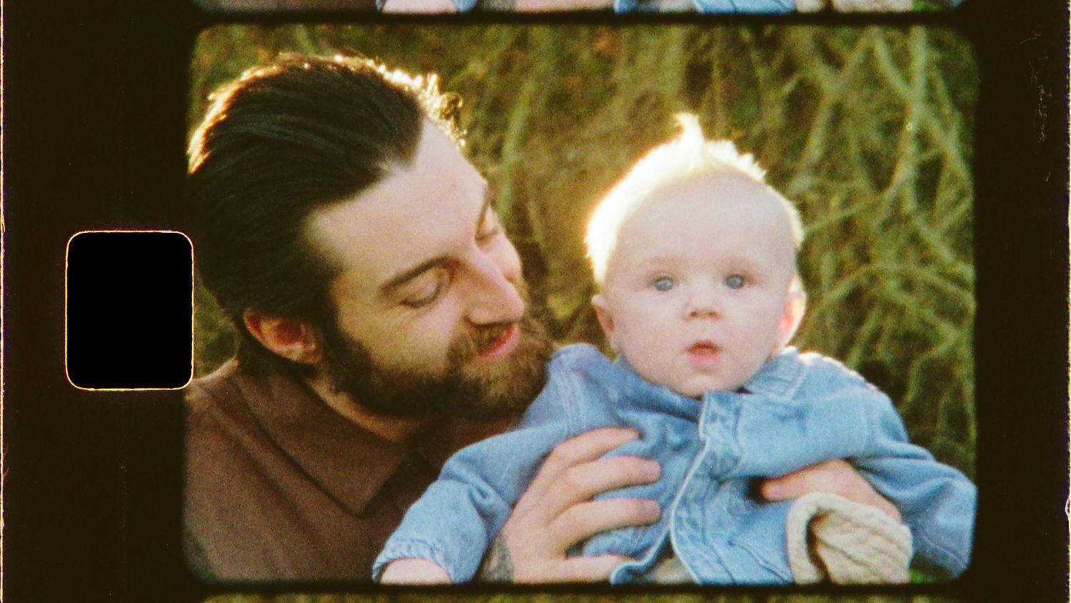 A bearded man tenderly holds a blue-eyed baby in a vintage film strip style photo outdoors.