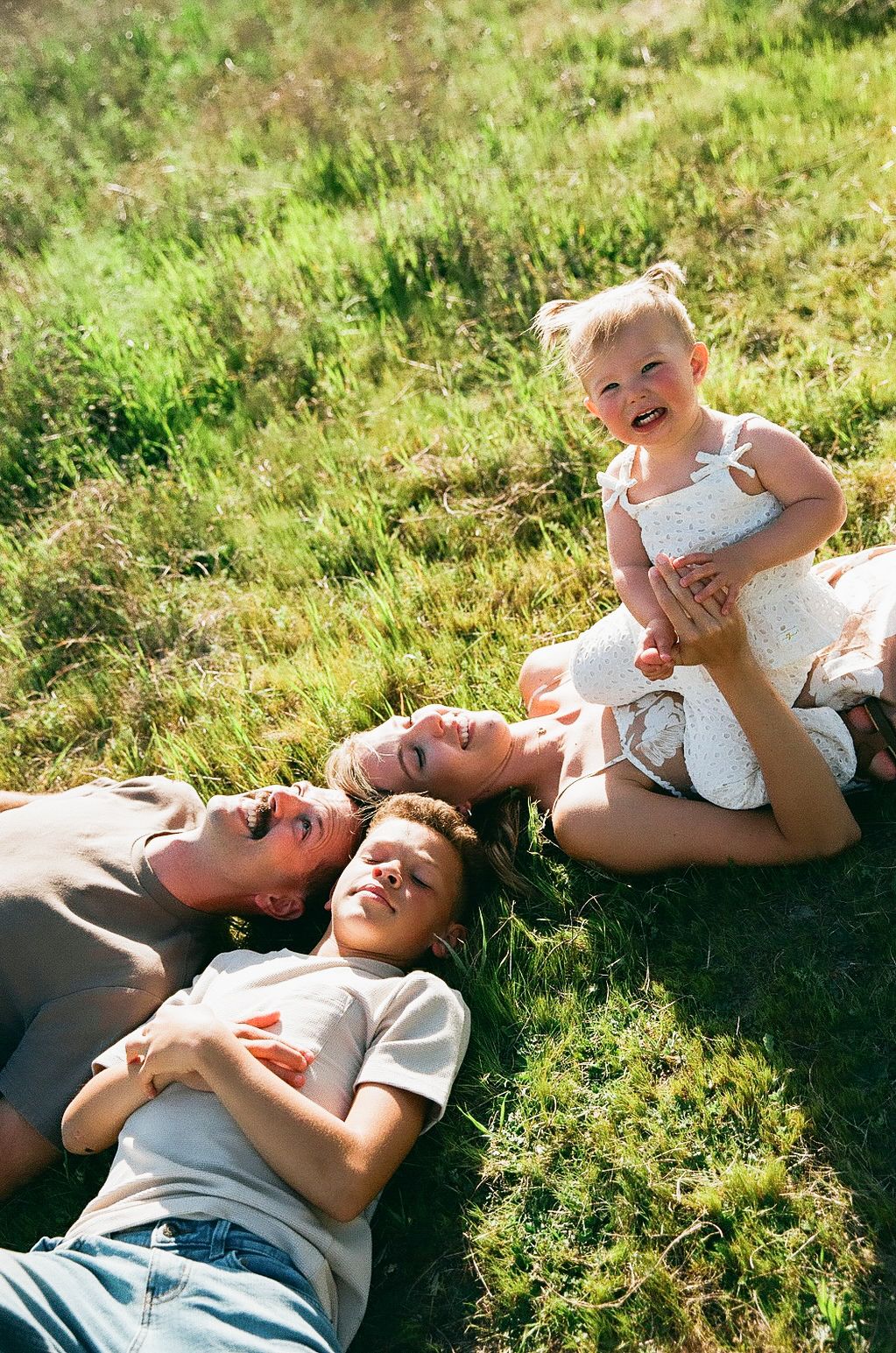 Happy family lies on sunny green grass while smiling toddler girl sits up in sunlight.