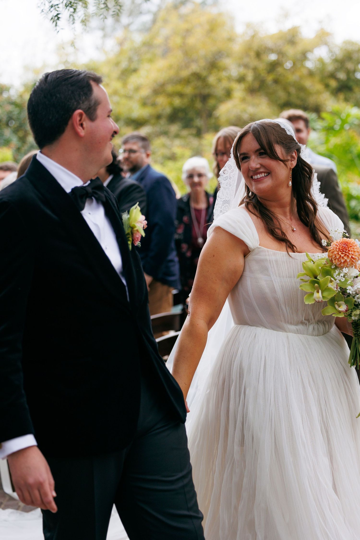 Bride in white tulle gown smiles at groom in black tuxedo during outdoor wedding ceremony.