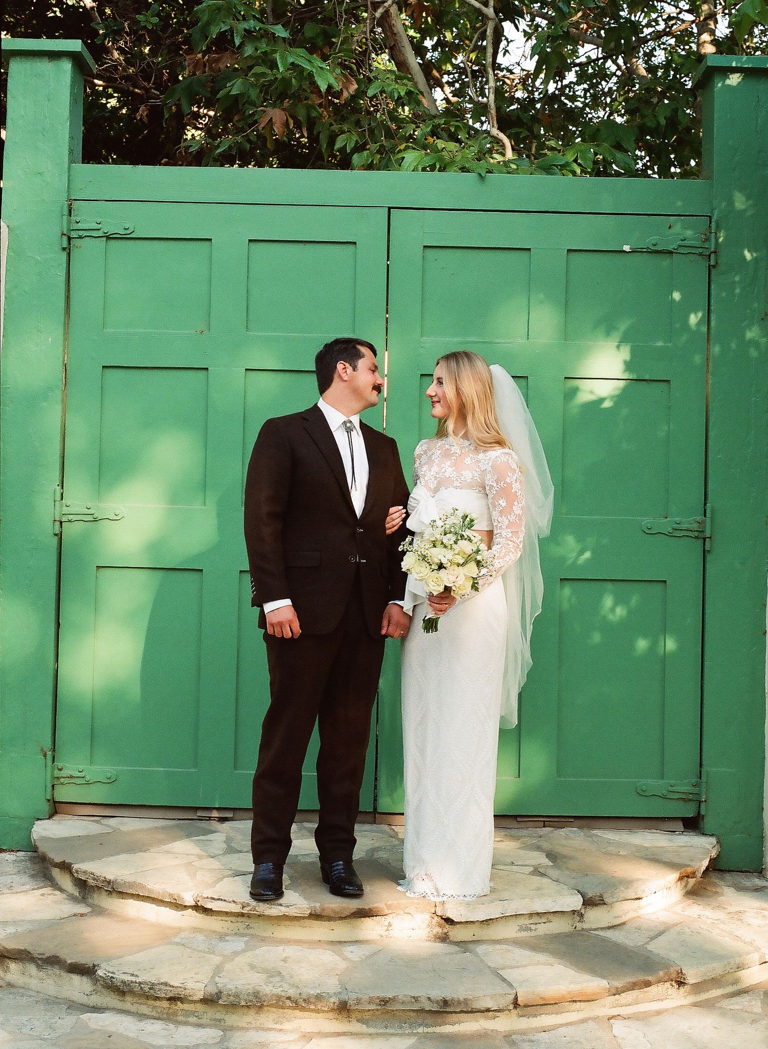 Bride and groom stand before a rustic green gate, smiling at each other, bride holding a white bouquet.