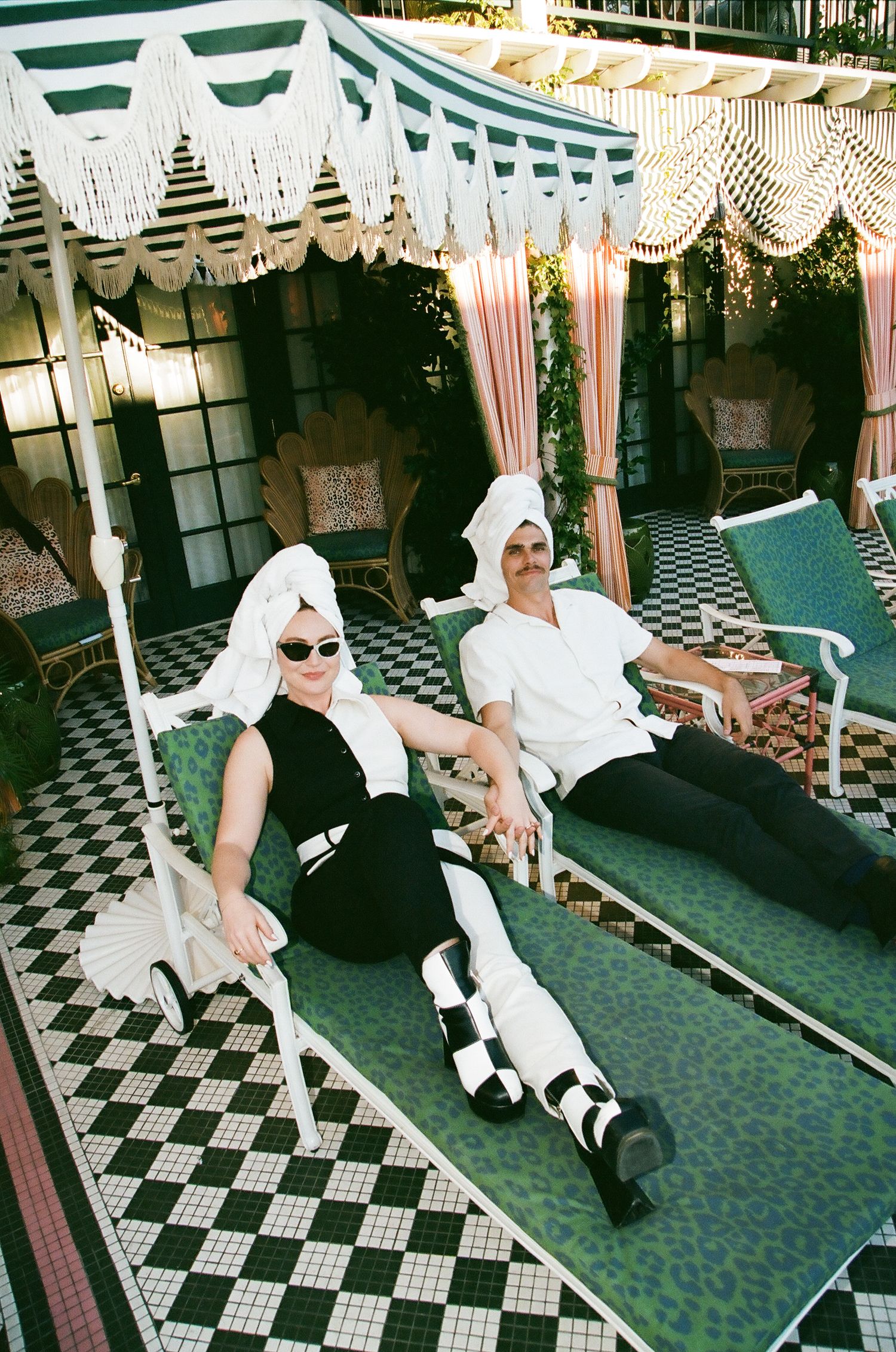 Couple lounging on outdoor chaise chairs with towels on heads at elegant black and white tiled patio.