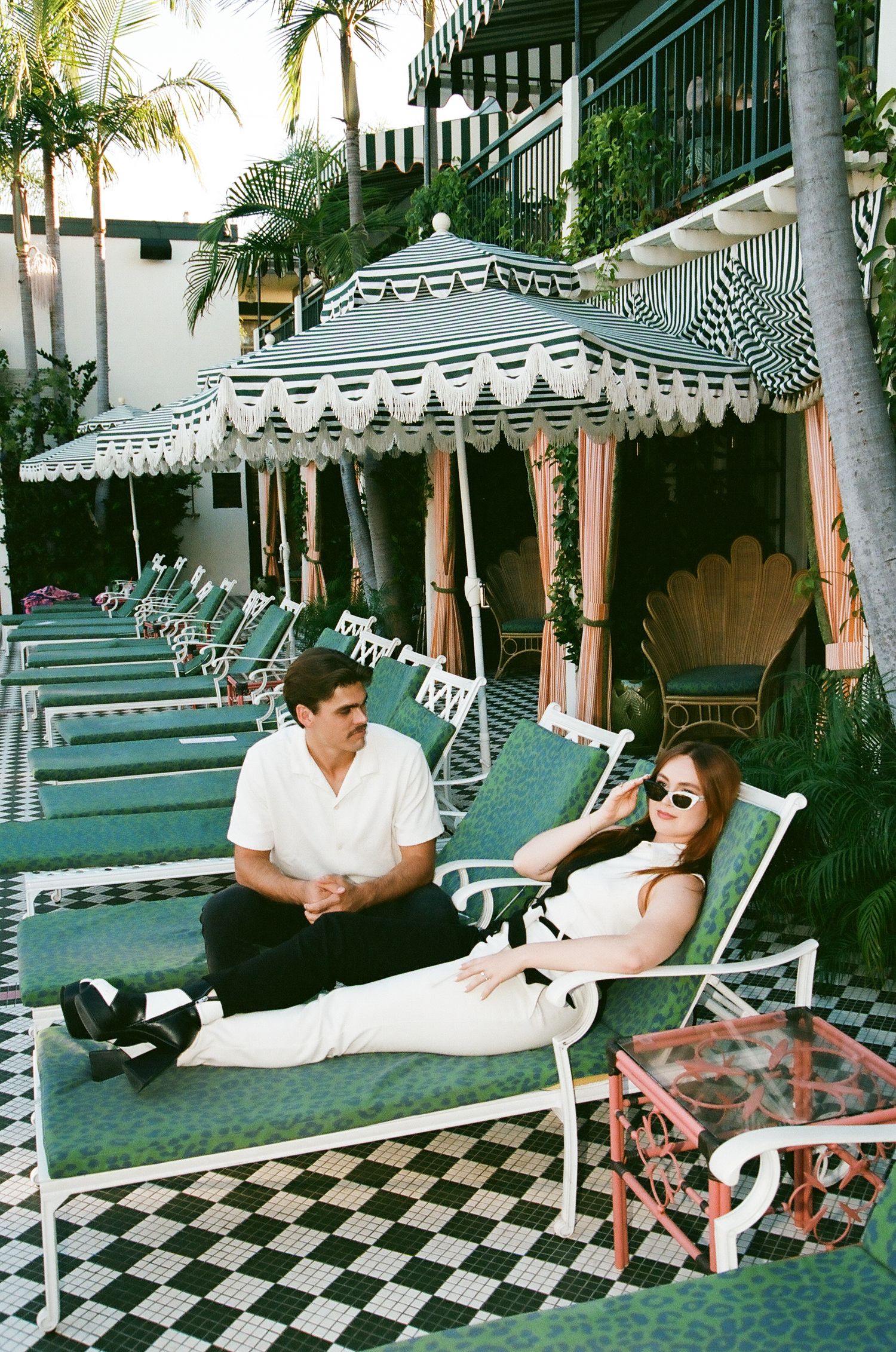 Couple relaxing by poolside on green lounge chairs under striped umbrellas at a luxury hotel.