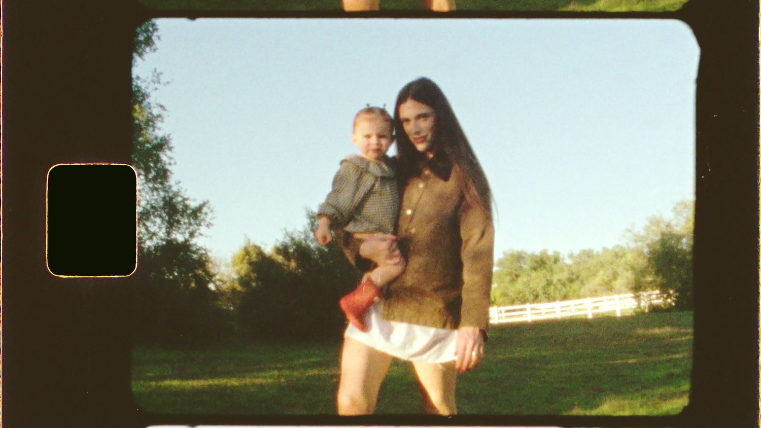 A woman holding a toddler poses outdoors on green grass with trees in the background, captured in vintage film style.