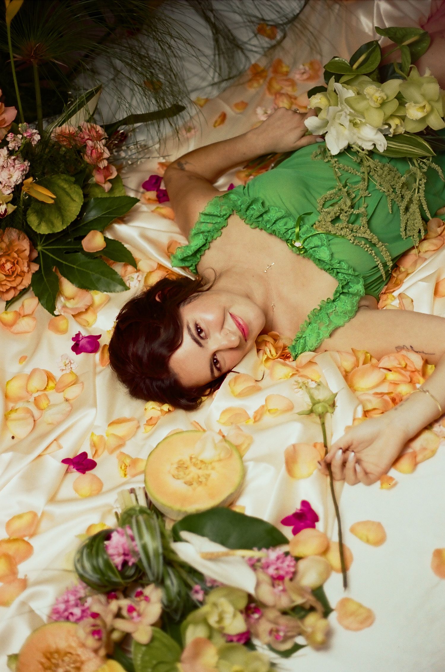 Woman in green top lying among flowers and rose petals with a cantaloupe melon on white satin fabric.
