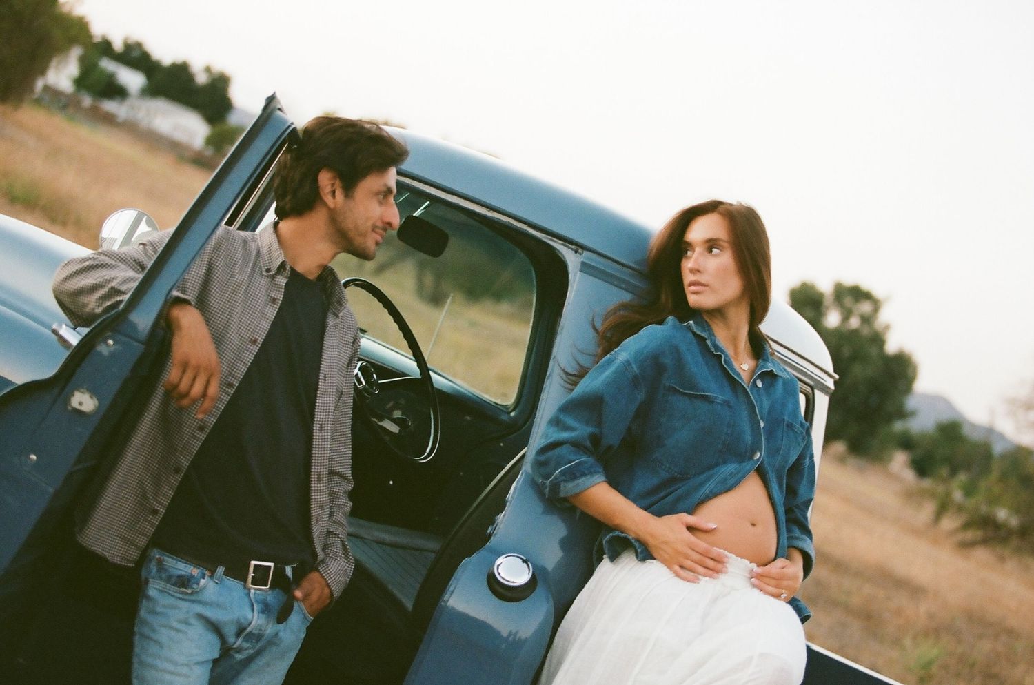 Pregnant woman in denim shirt and white skirt stands by vintage blue truck with man.