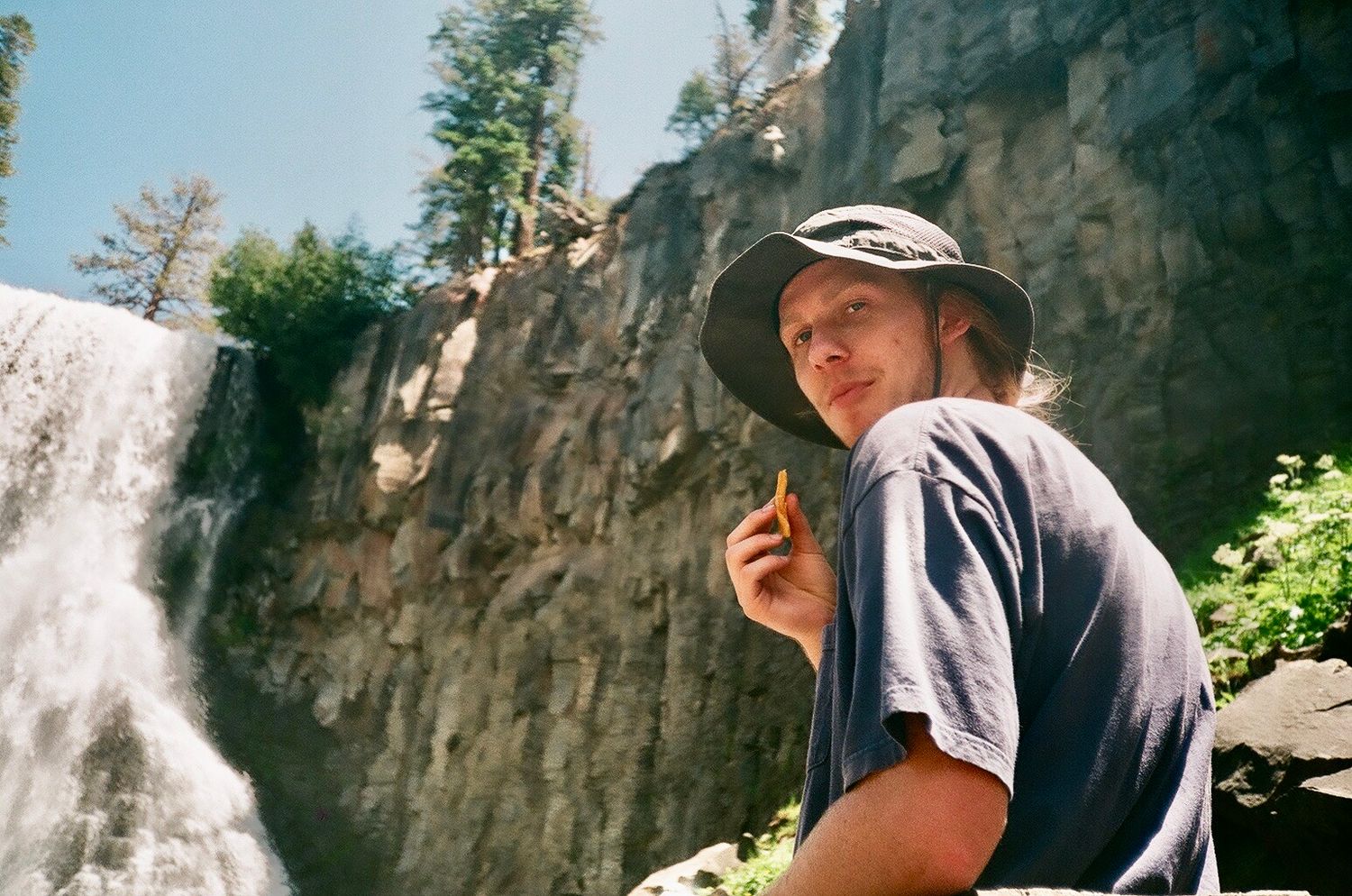 Young man in a bucket hat sitting by a waterfall on a sunny day in the mountains.