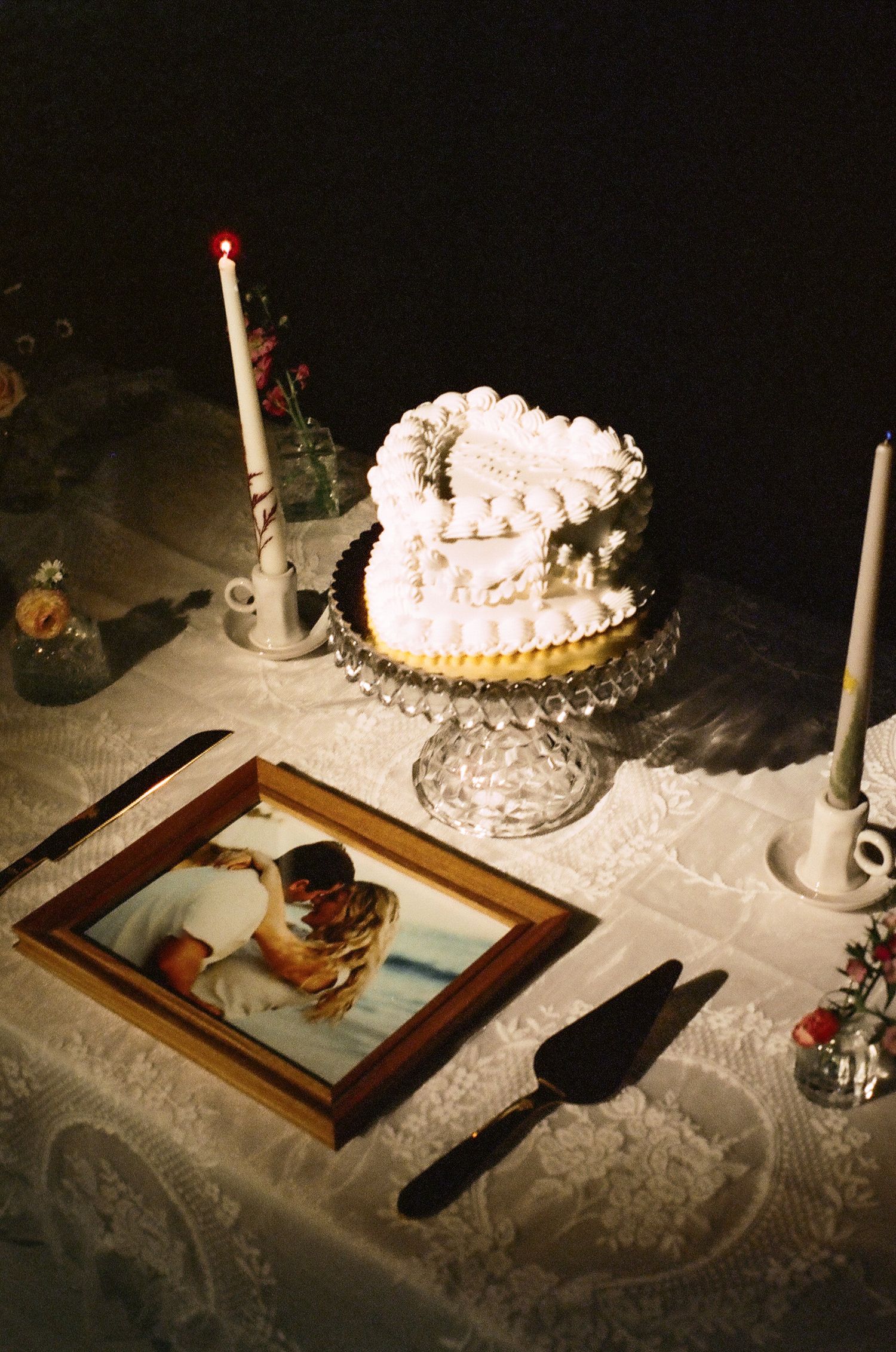 Heart-shaped wedding cake on crystal stand with candles and framed couple photo on lace tablecloth.