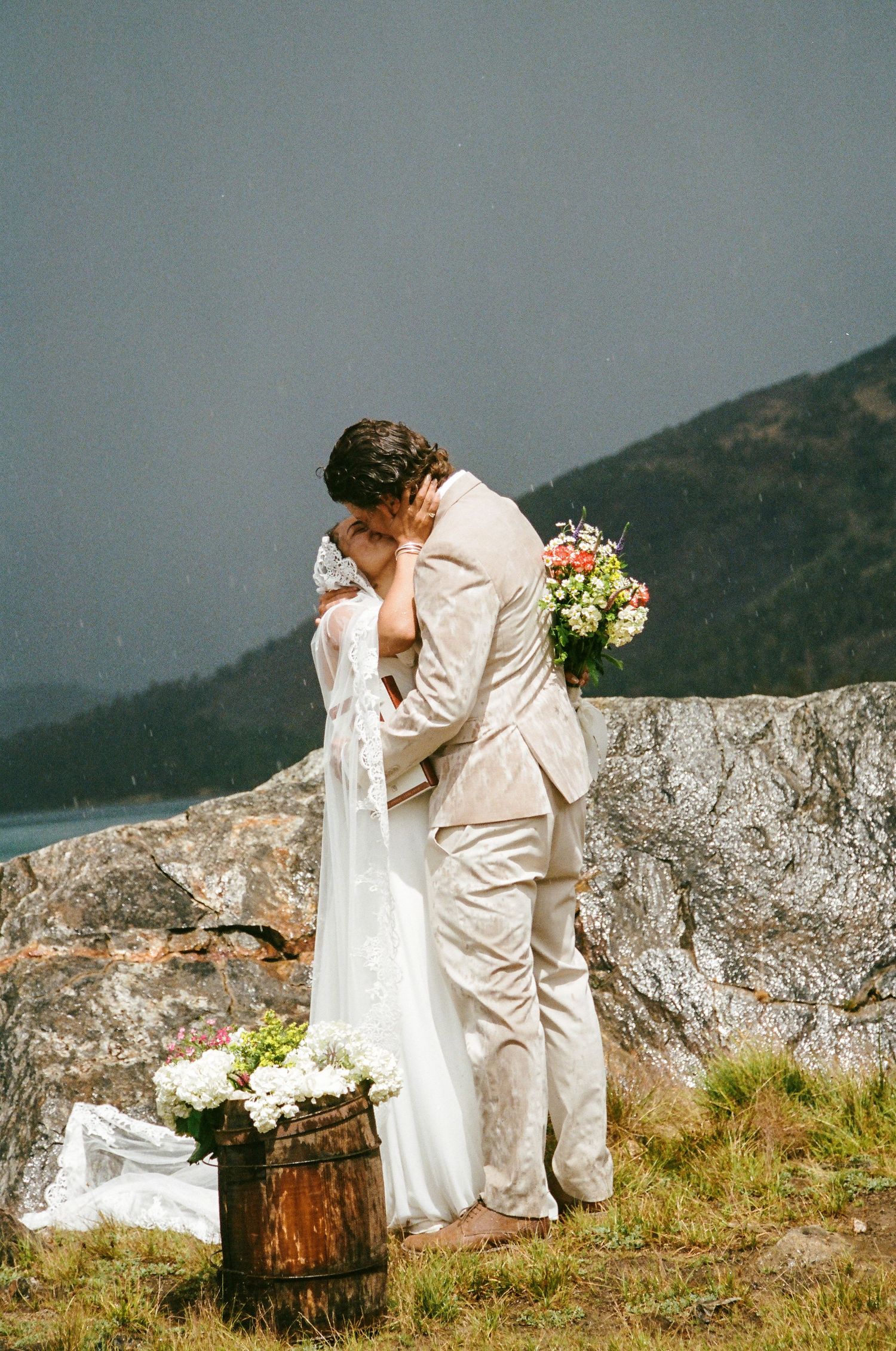 Bride and groom share a kiss on rocky mountain overlook, groom in tan suit holding bouquet, lake and hills in background.