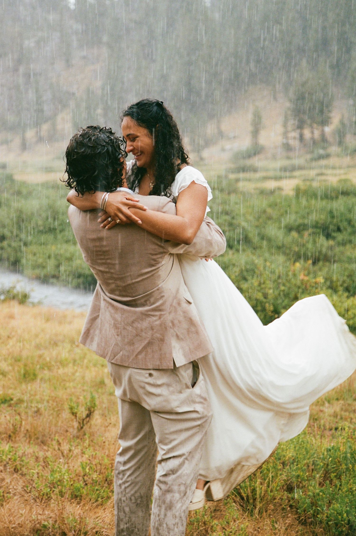 Groom lifts bride in white dress during romantic rainy outdoor wedding ceremony in a lush green meadow.