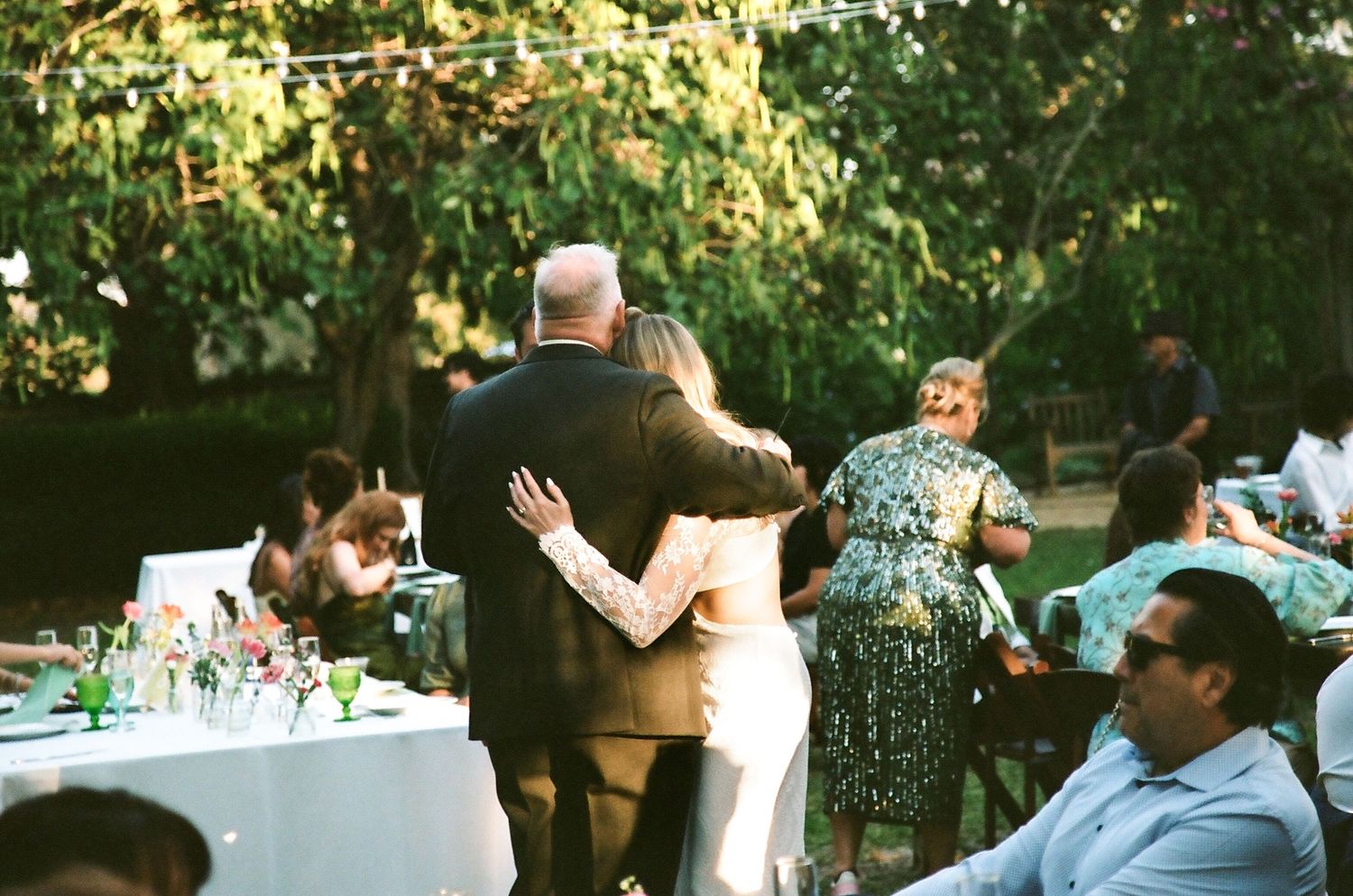 Couple sharing a warm embrace at an outdoor wedding reception surrounded by guests and decorated tables.