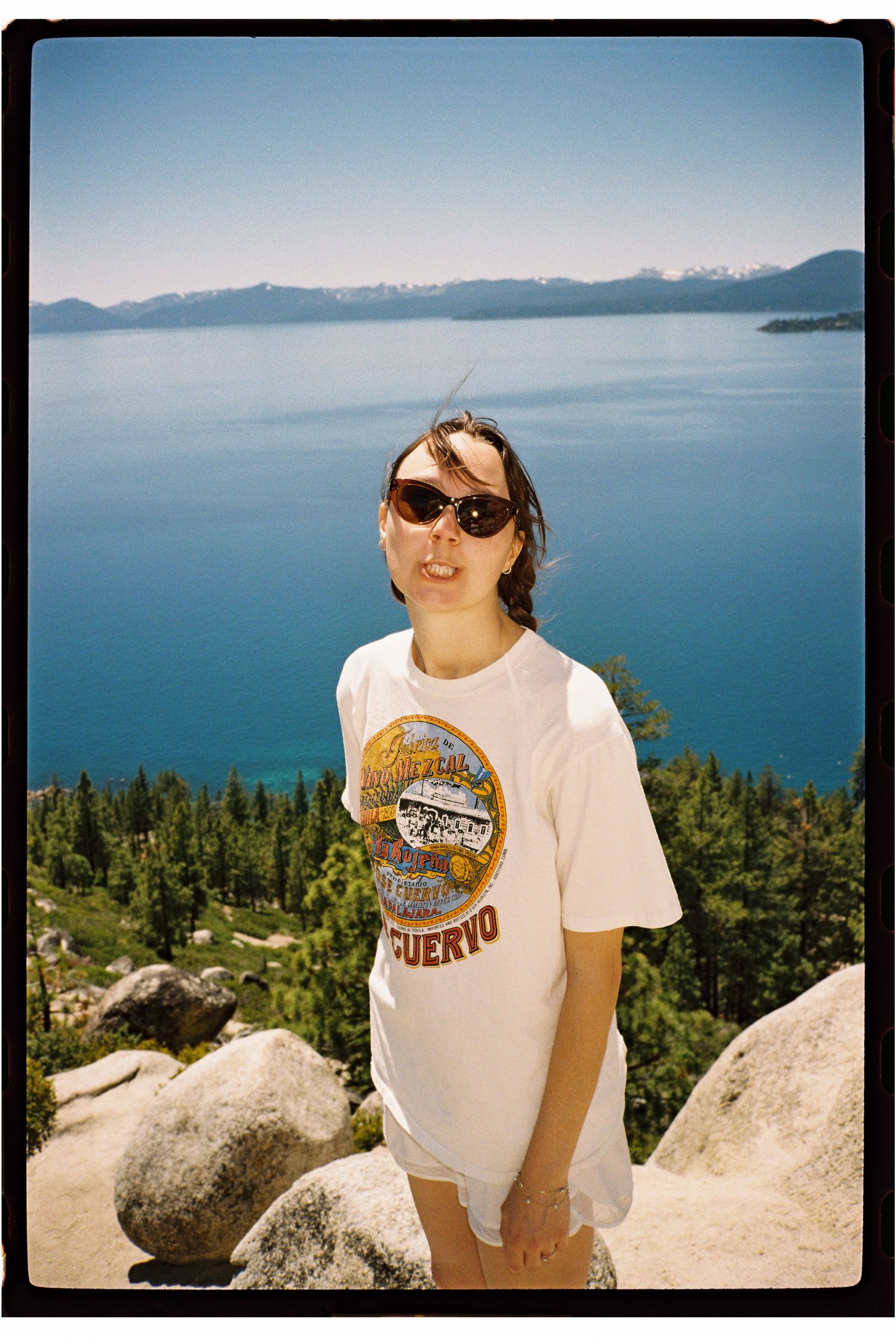 Woman in Jose Cuervo tee and sunglasses poses at Lake Tahoe overlook with blue water and mountains.