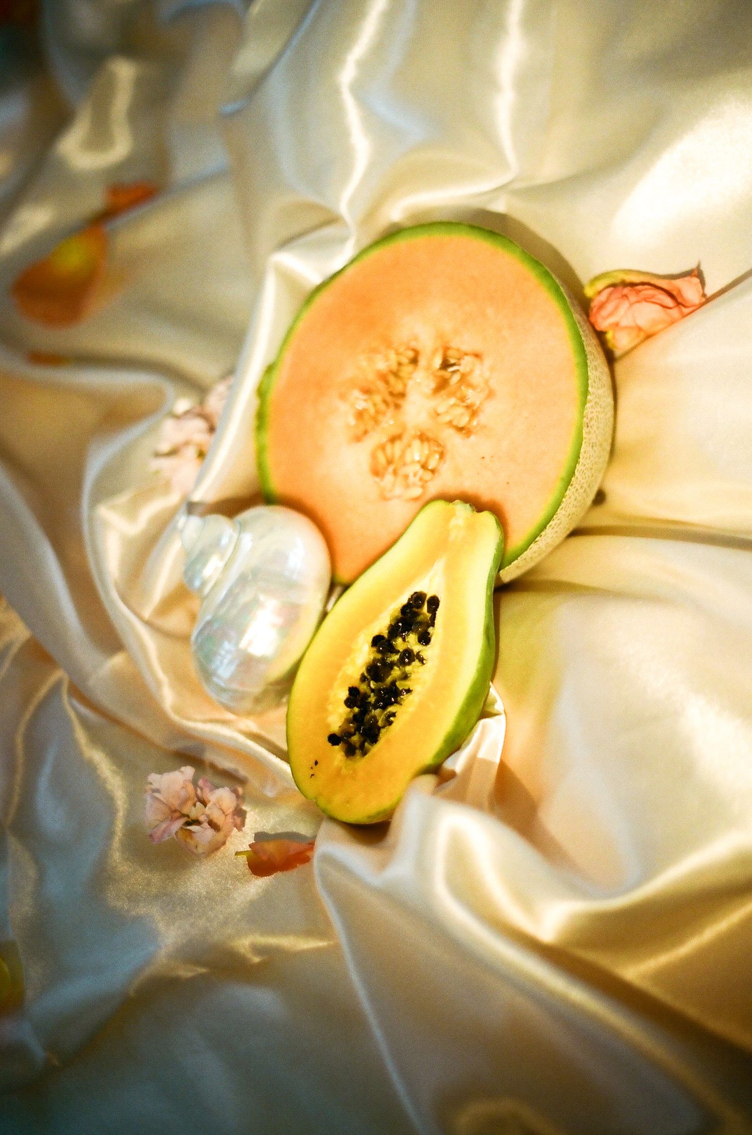 Halved cantaloupe and papaya with pearl beads on luxurious white satin fabric in warm sunlight.