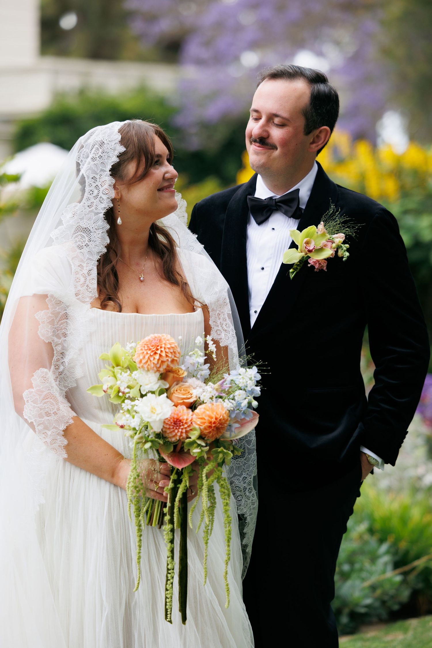 A bride in a white dress with lace veil holds a dahlia bouquet while smiling at her mustachioed groom in a black tuxedo.