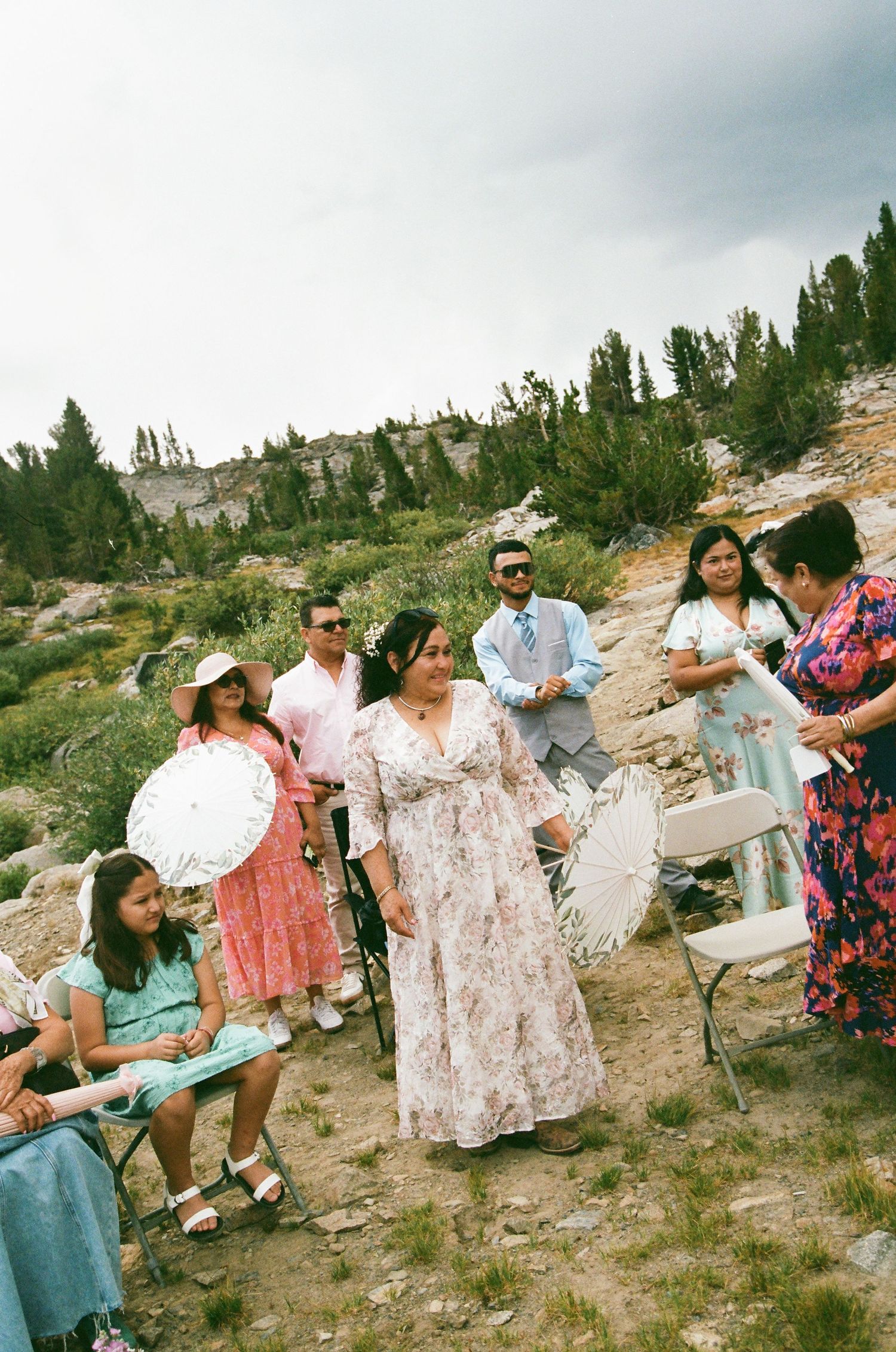 Outdoor wedding ceremony with guests in floral dresses holding parasols on a rocky mountain landscape.