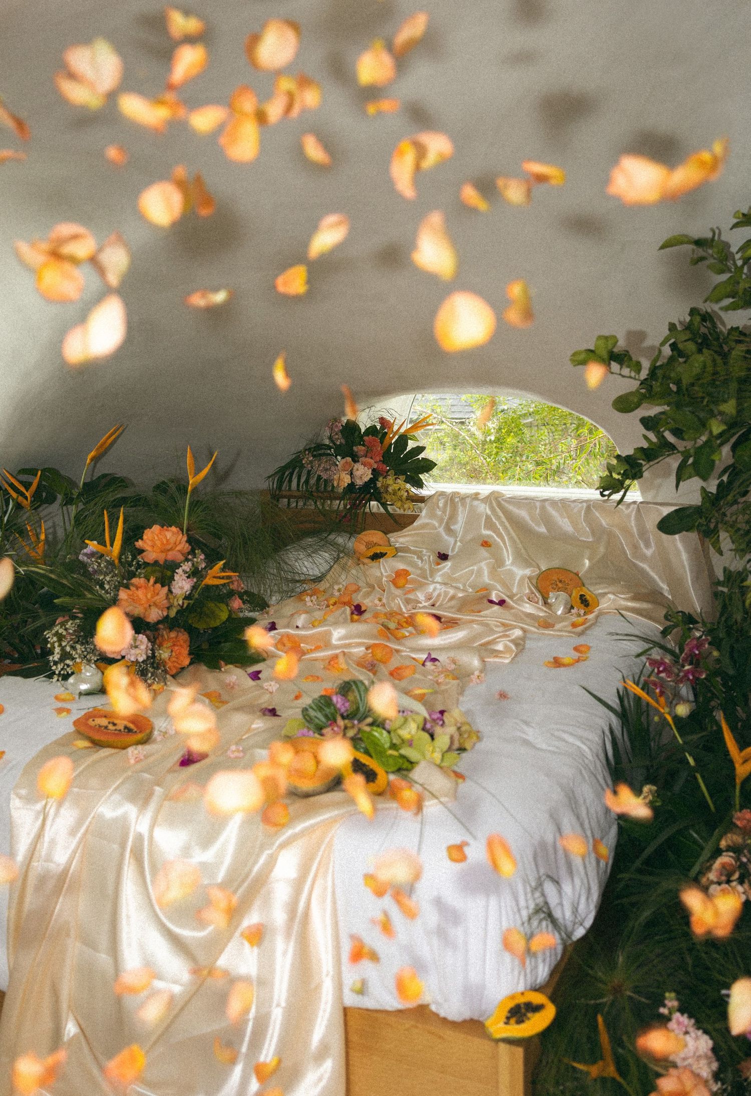 Orange rose petals floating above a white satin bed with tropical flowers in a curved white room.