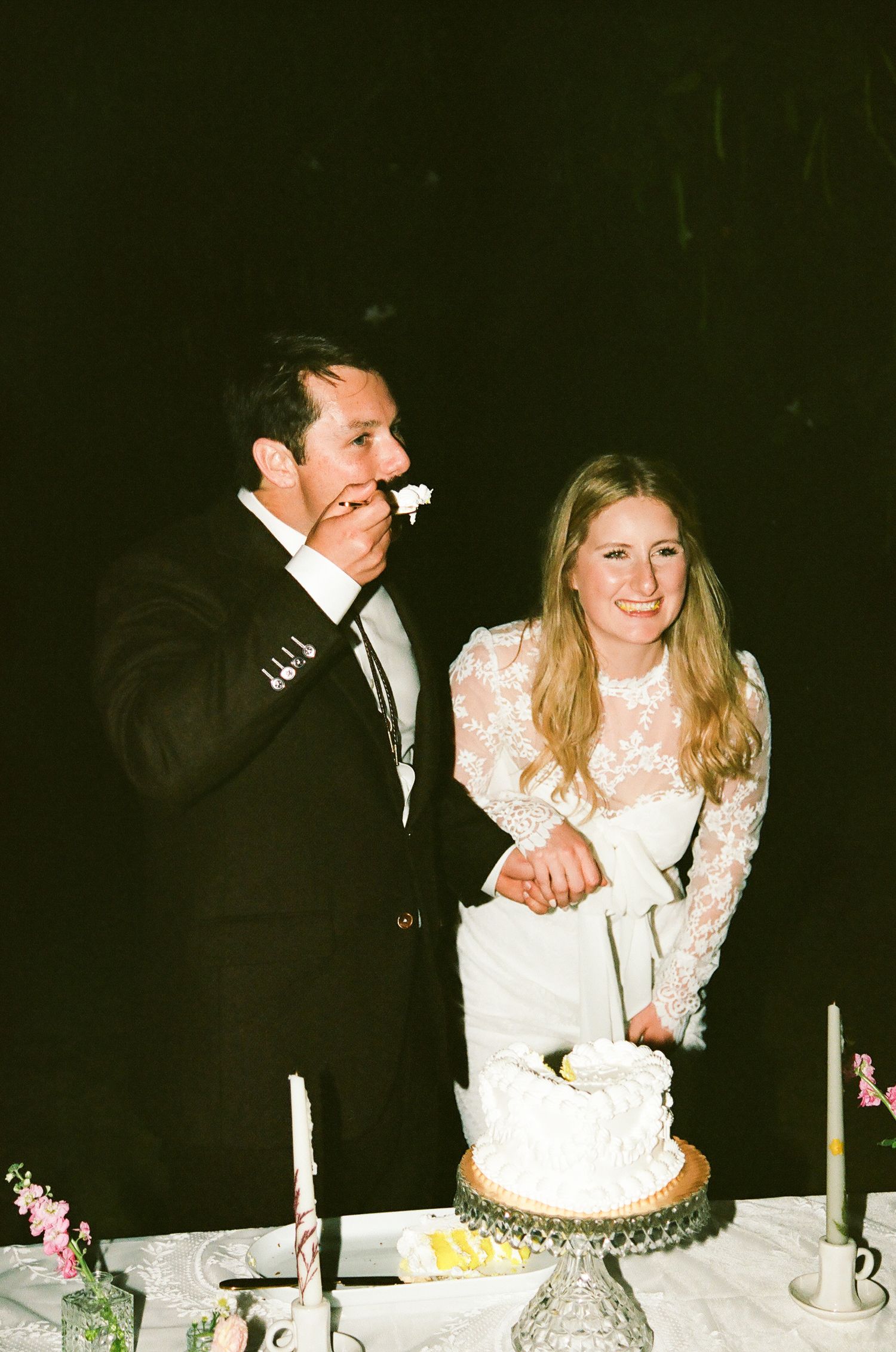 Bride and groom cutting white wedding cake at reception table with candles, groom laughing joyfully.